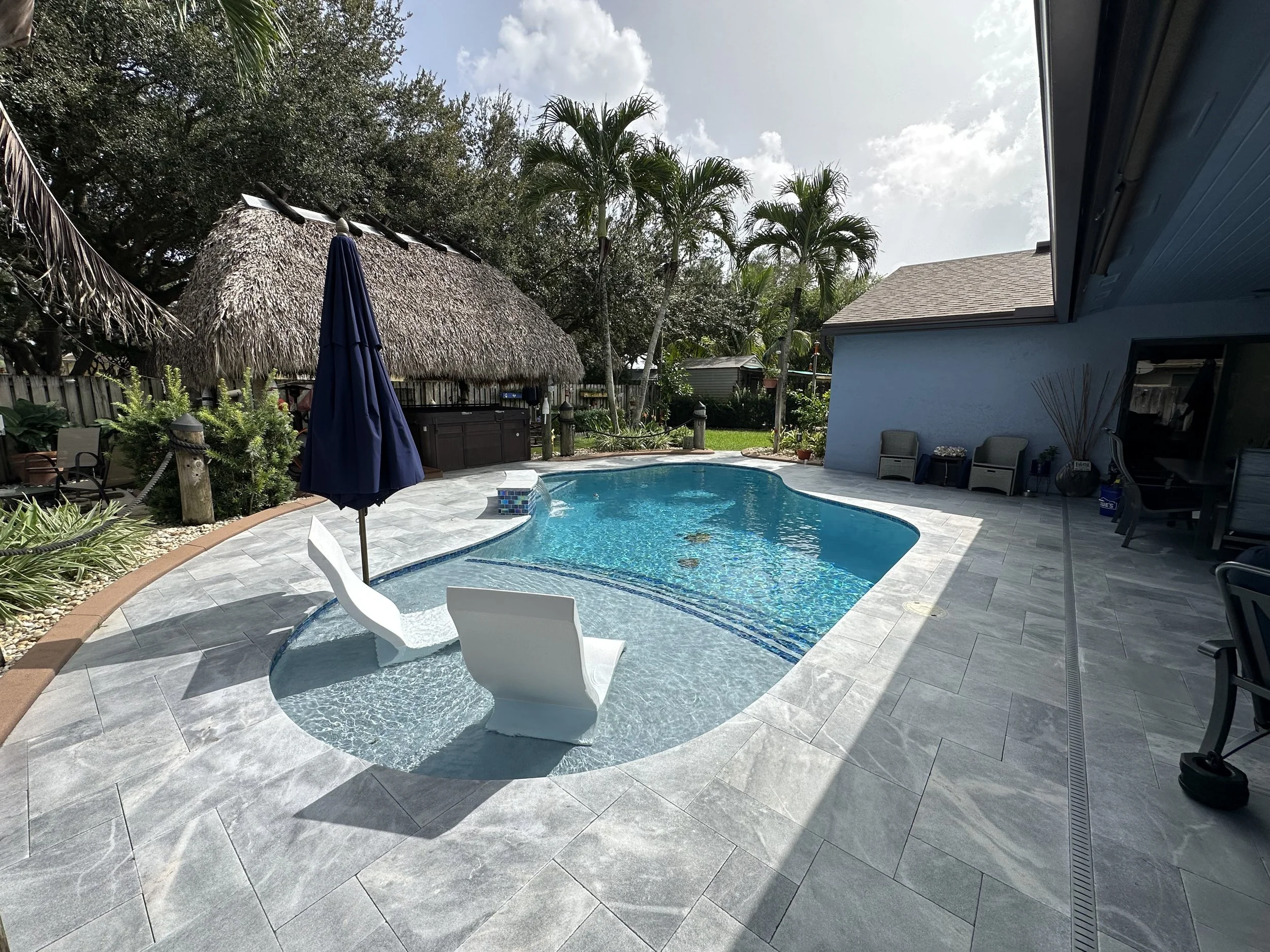 Backyard pool with sunken seating, umbrella, and tropical landscaping including palm trees and a tiki hut.
