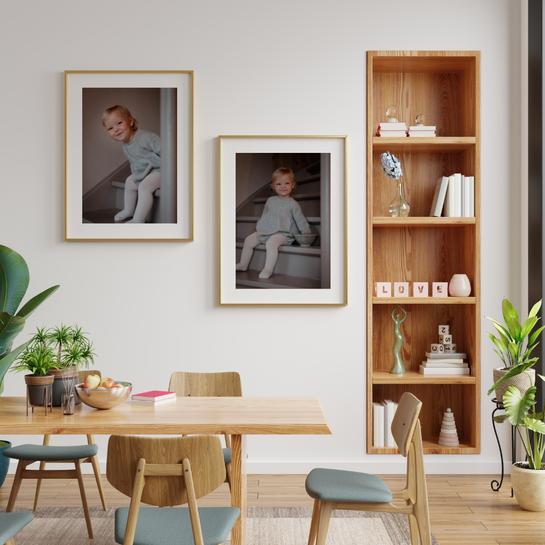 Innendørs bilde av en spisestue med tre bilder av en liten jente på veggen, en trehylle med dekorasjoner og bøker, og grønne planter på bord og gulv.