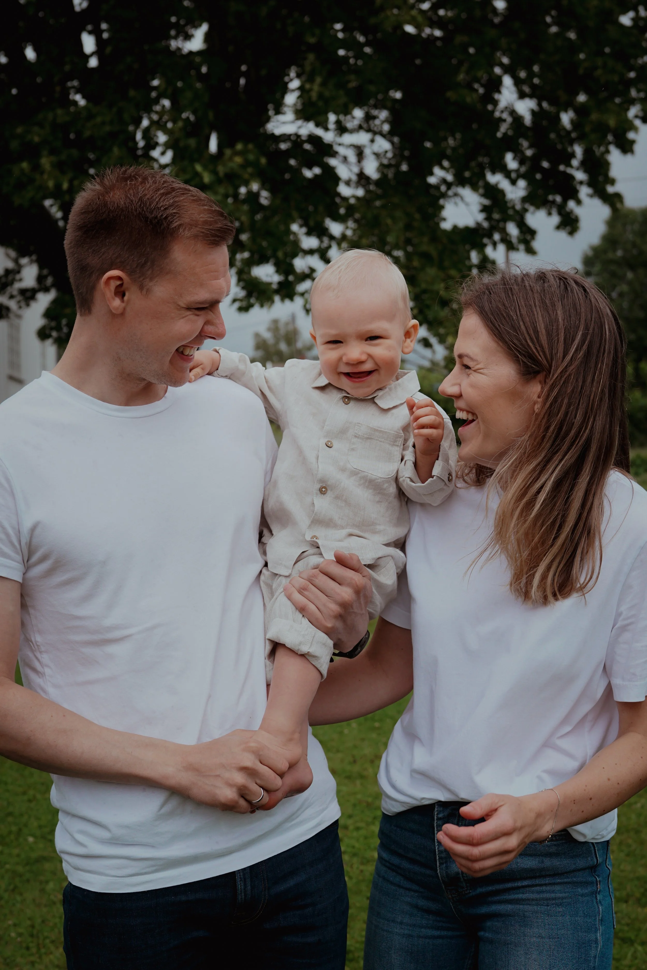 Et lykkelig familie med to voksne og ett barn ler sammen ute i en park.