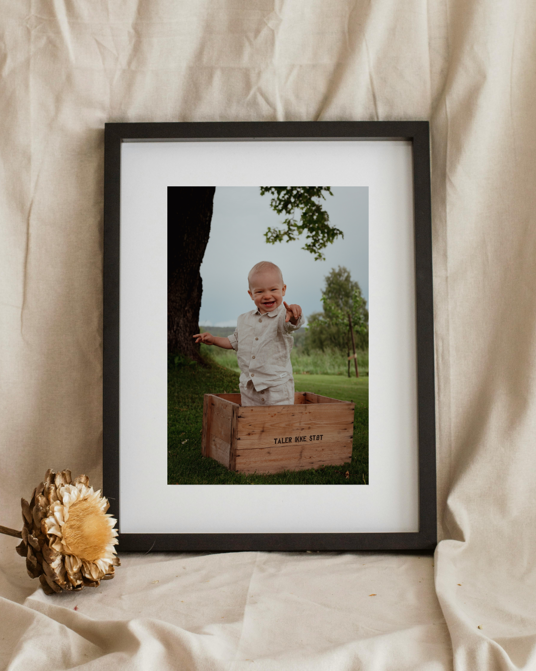 Barnebilde av en liten gutt som står i en trekasse i en park, smiler og peker mot kamera.