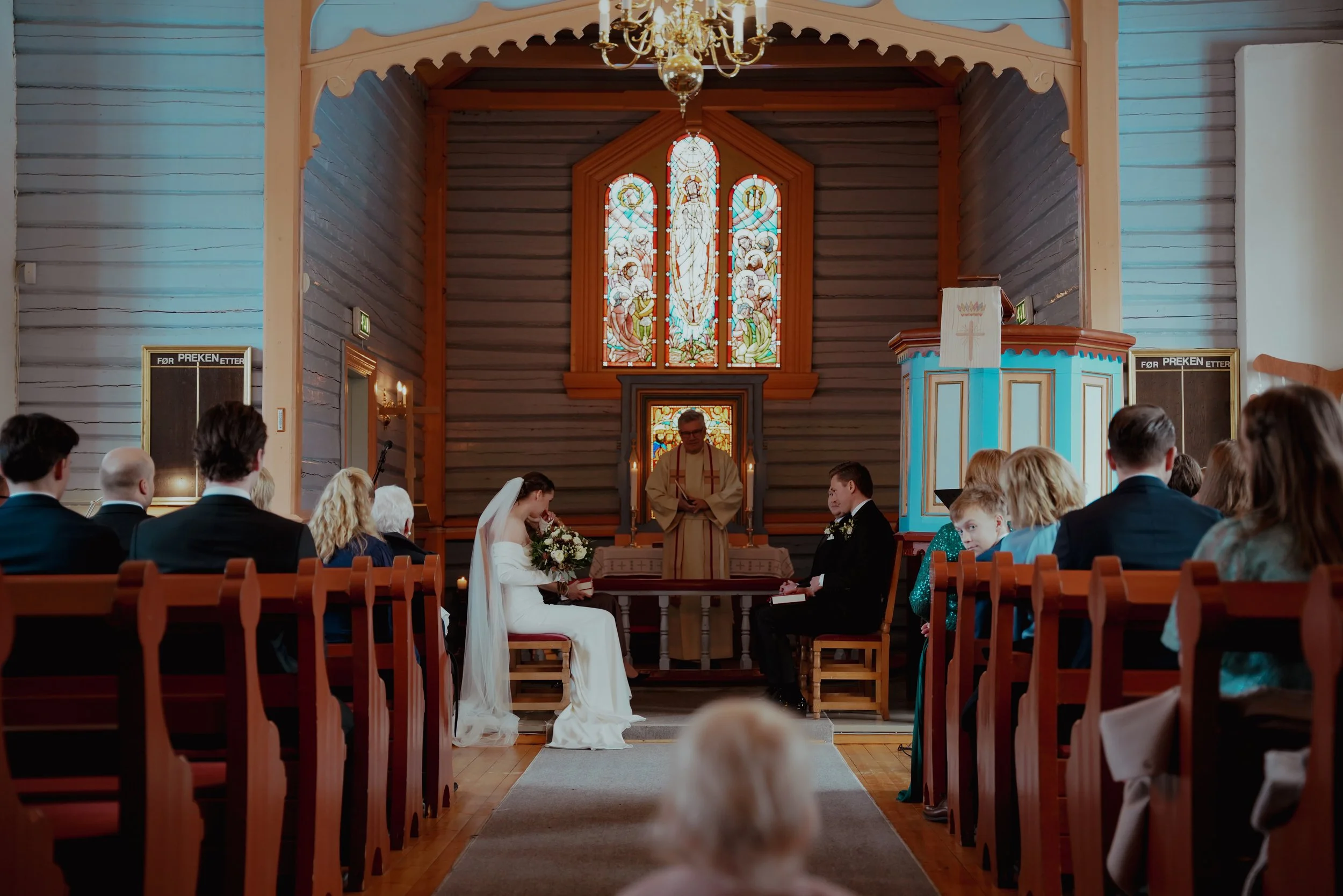 Bryllup inne i en kirke med brud og brudgom foran alteret, omgitt av gjester, under en dekorativ lyskrone og et farget glassmaleri i bakgrunnen.