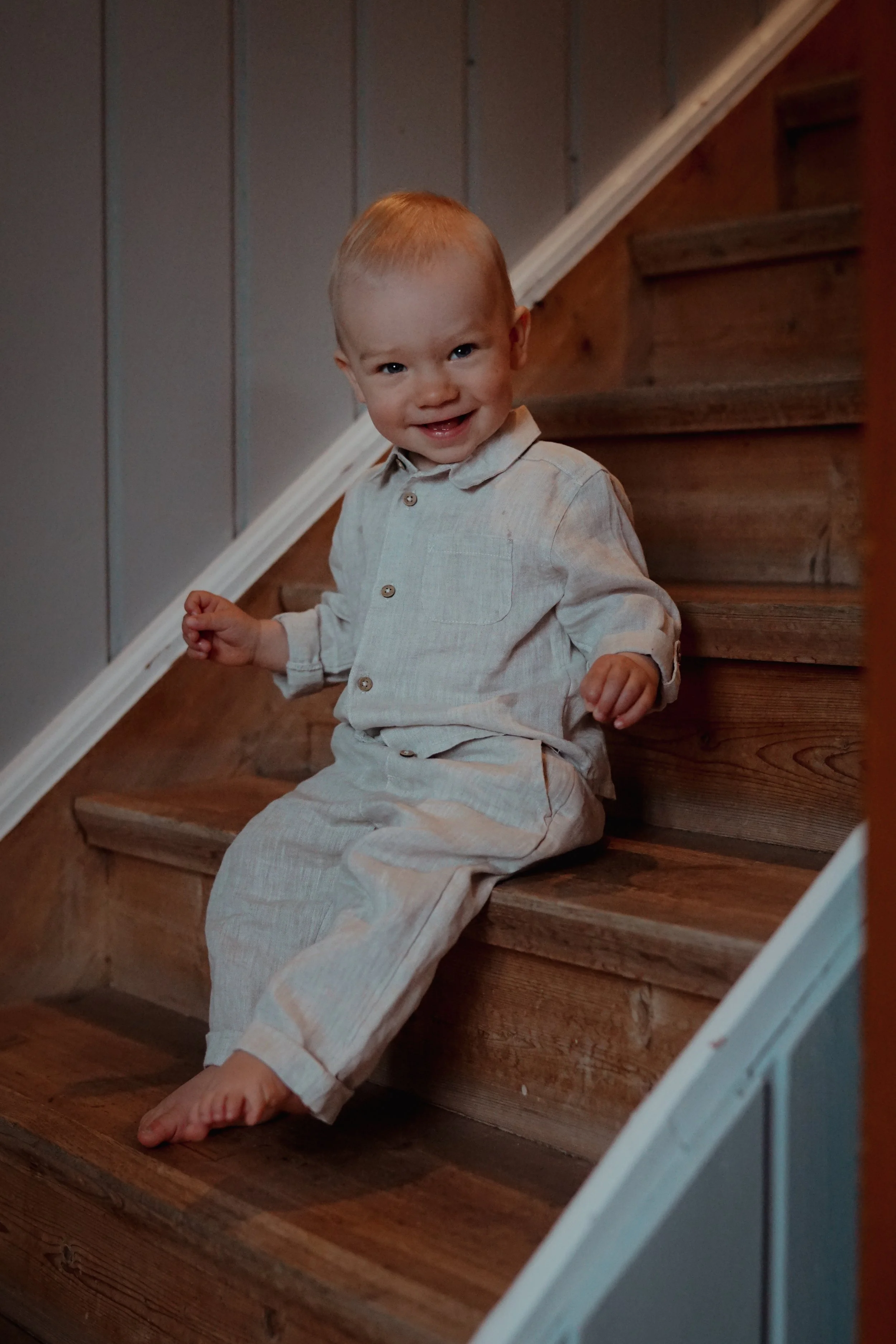 Liten gutt som sitter på trappen, smilende og ser mot kamera, iført lyst klær.
