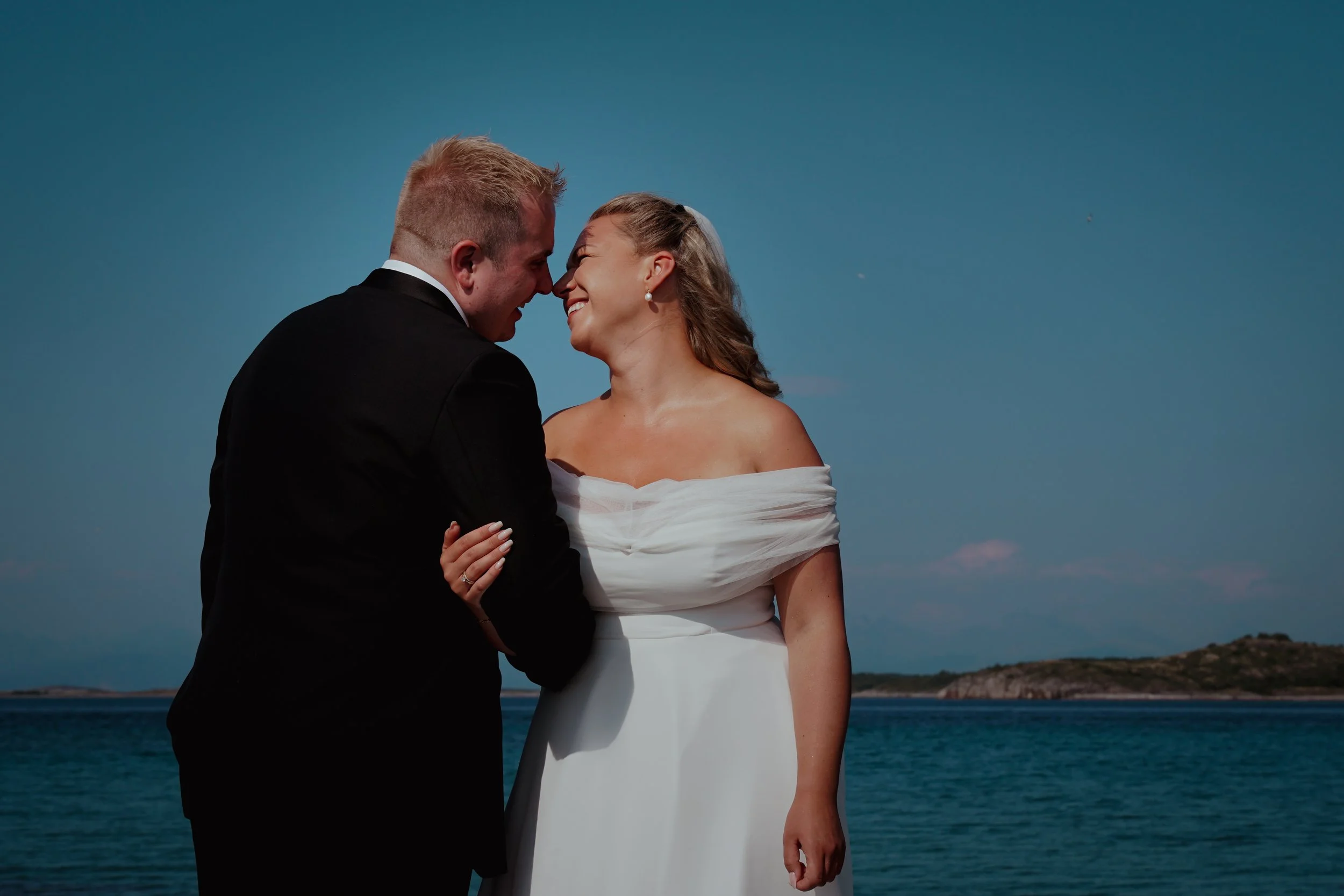 Et lykkelig par som står tett sammen, smiler og ler, foran sjøen og en klar himmel.