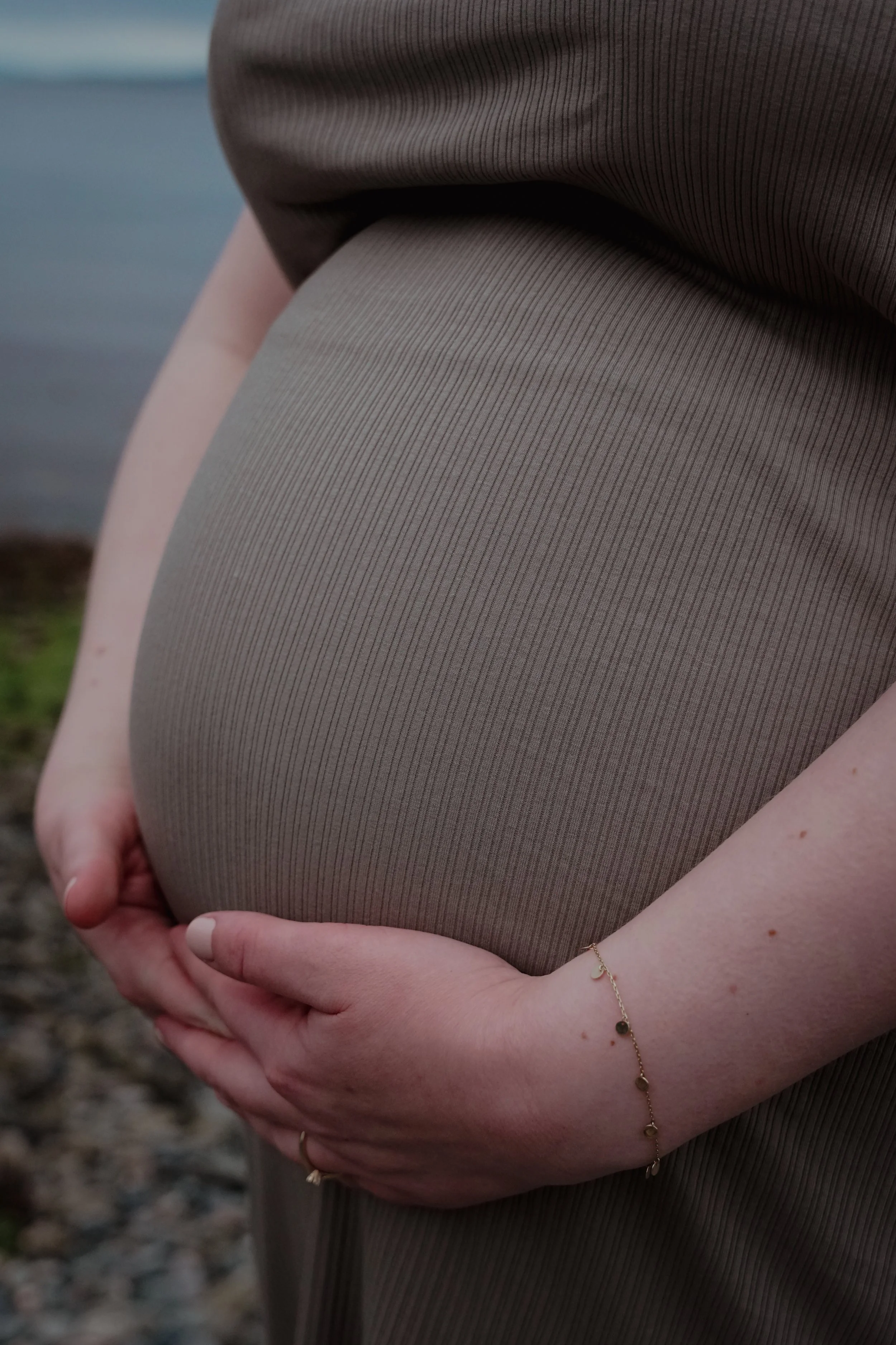 Nærbilde av en gravid persons mage som holdes av begge hender, iført en grå kjole med tynne striper og en tynn armbånd.