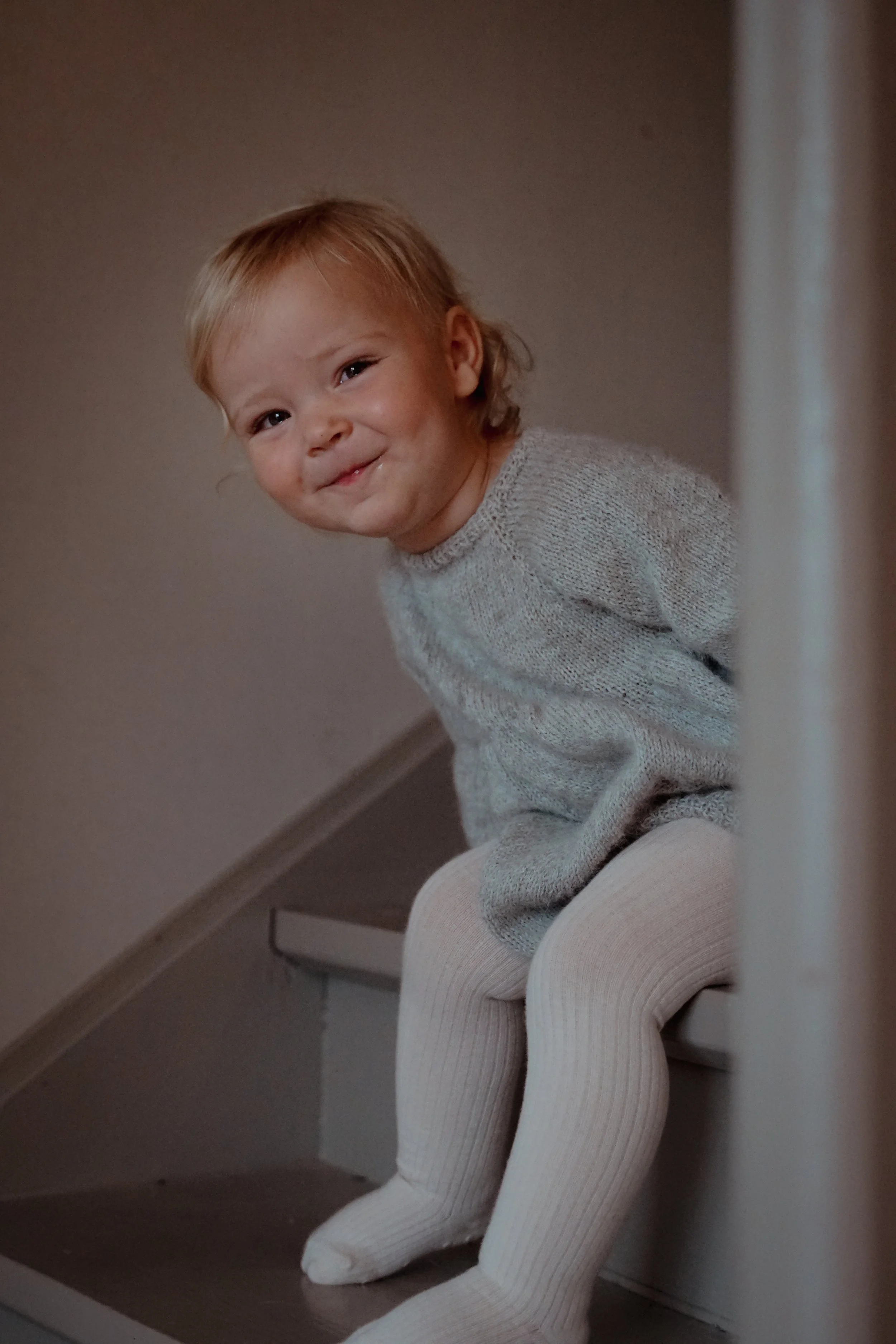 Liten gutt med blond hår smilende, sitter på en trapp, iført lyse tights og grå genser.