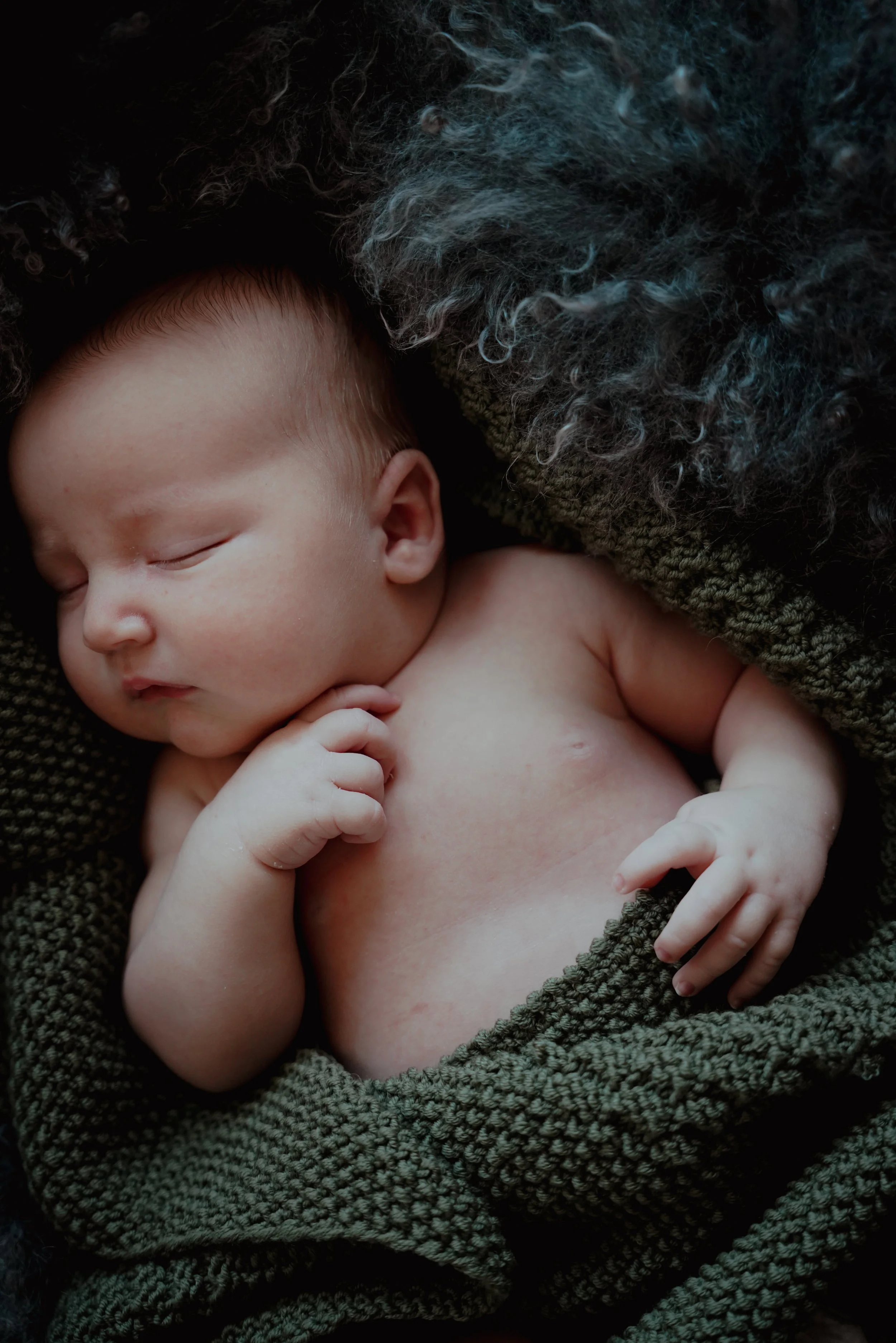 Sovende baby ligger på et mykt, mørkt grønt teppe med et saueskinn under seg.