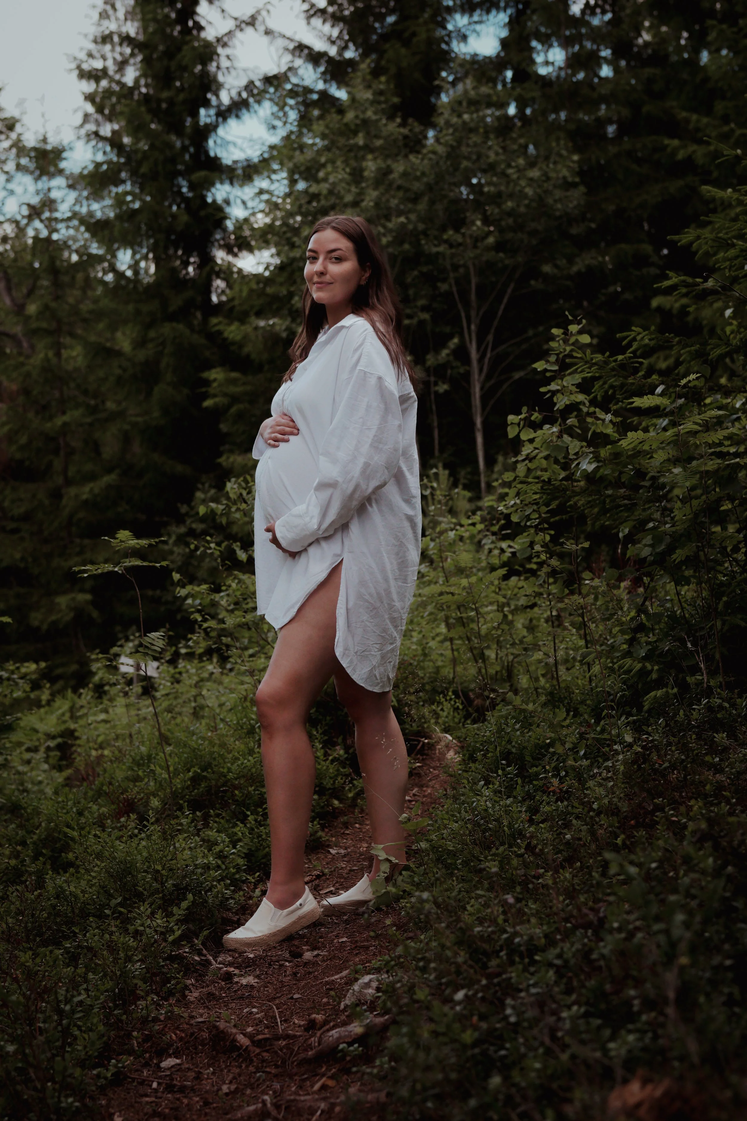 Gravid kvinne i en hvit skjorte og hvite sneakers går på en sti gjennom skogen, omgitt av grønne trær og busker.