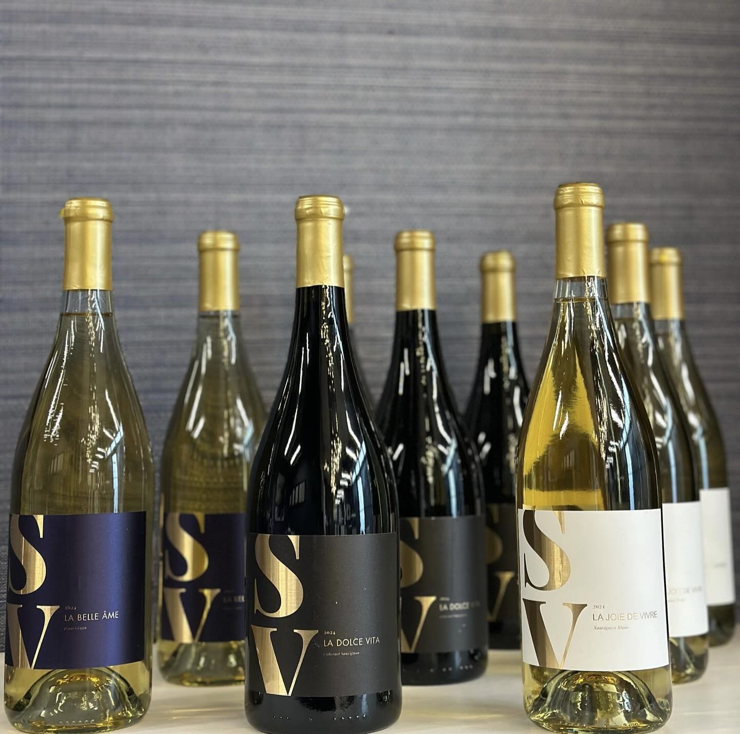 Assorted bottles of wine with SS and 'La Belle Âme, La Dolce Vita, La Joie de Vivre' labels and gold caps on a light background.