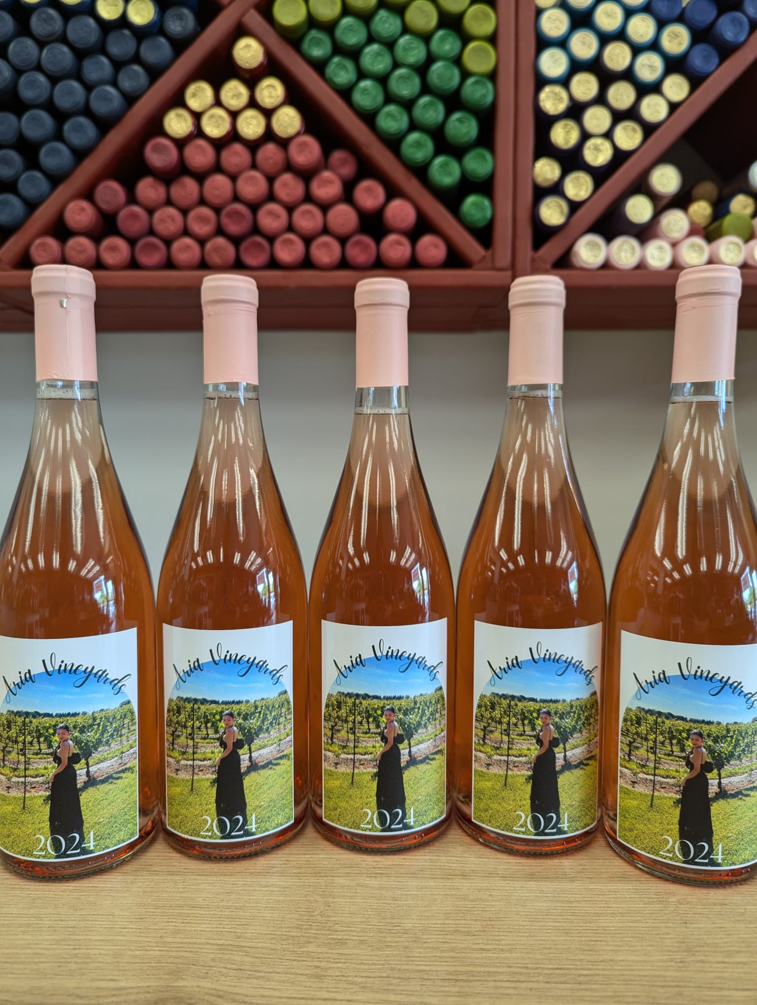 Five bottles of Aria Vineyards 2024 rosé wine with pink caps on a wooden shelf, set against a background of colorful wine bottle caps arranged on a triangular rack.