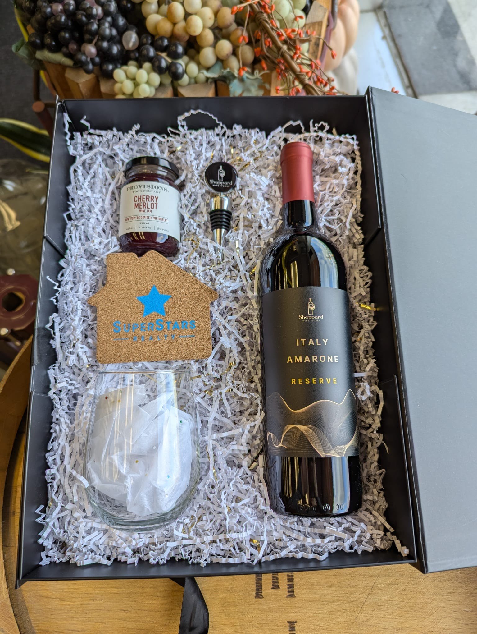 Gift box containing a wine bottle labeled 'Italy Amarone Reserve,' a jar of cherry Merlot jam, a cork and stopper, a star-shaped cork coaster labeled 'SuperStars,' and a wrapped wine glass, all arranged on shredded paper.