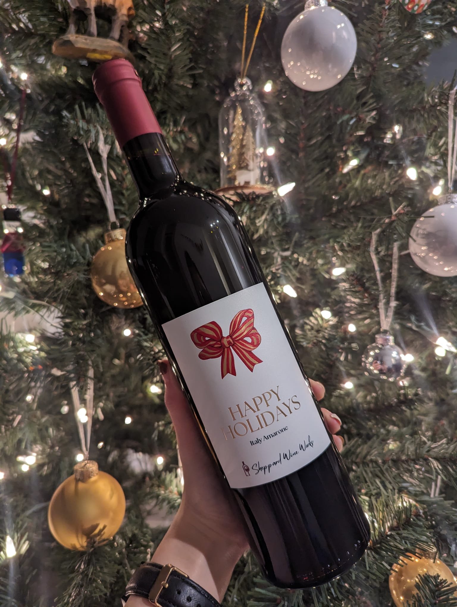 A person holding a bottle of wine with a holiday-themed label in front of a decorated Christmas tree, featuring ornaments and lights.