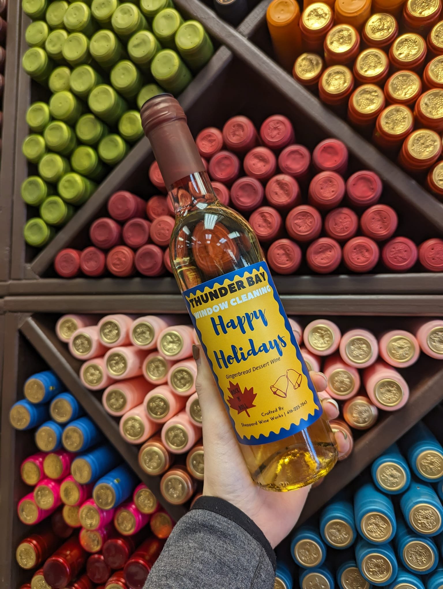 Person holding a bottle of gingerbread dessert wine labeled "Happy Holidays" against a background of colorful wine bottles stacked in diagonal rows.
