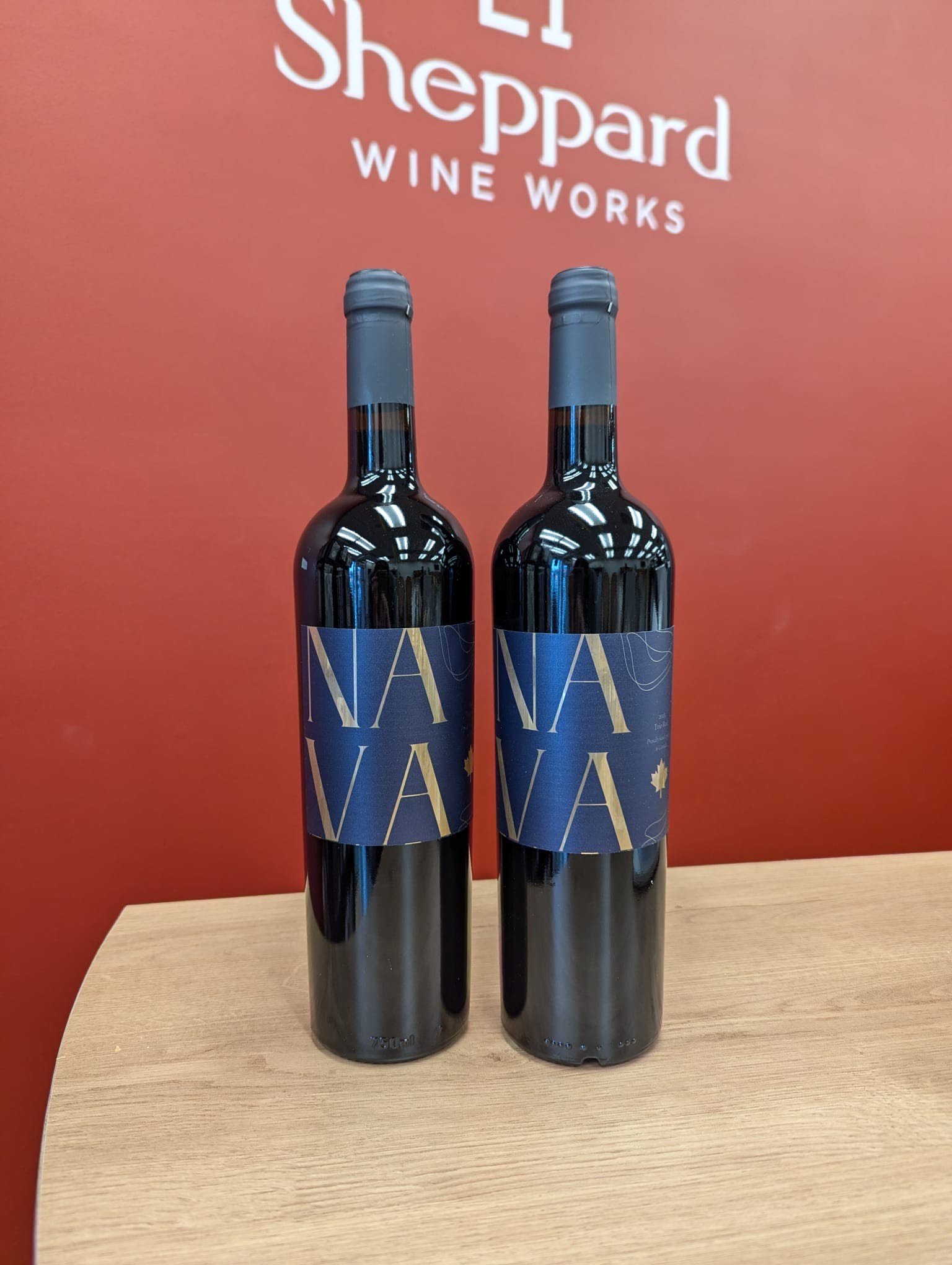 Two bottles of red wine with blue labels displaying 'NAVA,' set against a red wall with a 'Sheppard Wine Works' sign.