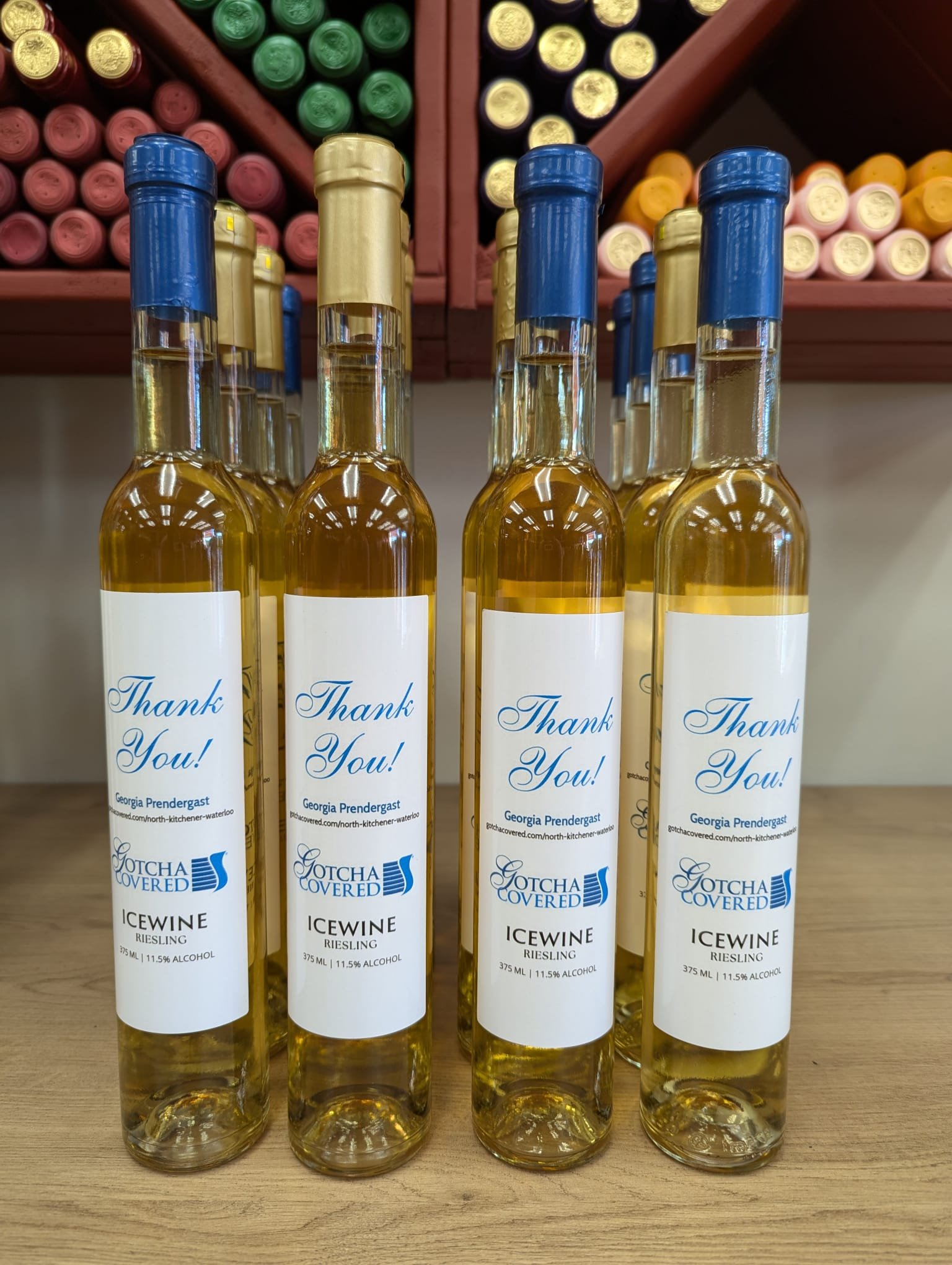 Bottles of Gotcha Covered Icewine Riesling with "Thank You!" labels displayed on a wooden shelf, surrounded by wine racks filled with various colored bottles.