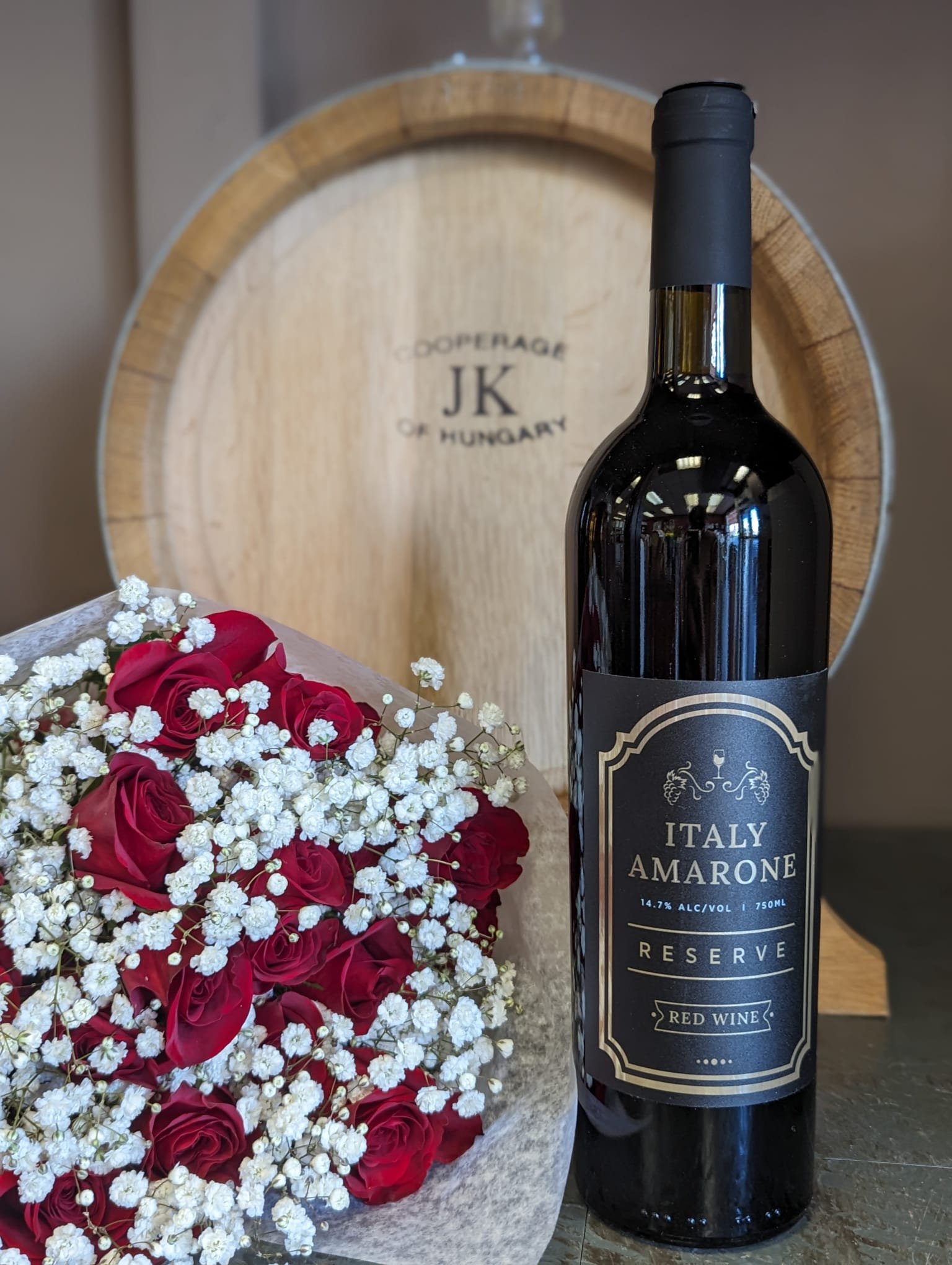 Bottle of Italy Amarone Reserve red wine beside a bouquet of red roses and white baby's breath, with a wooden wine barrel in the background.
