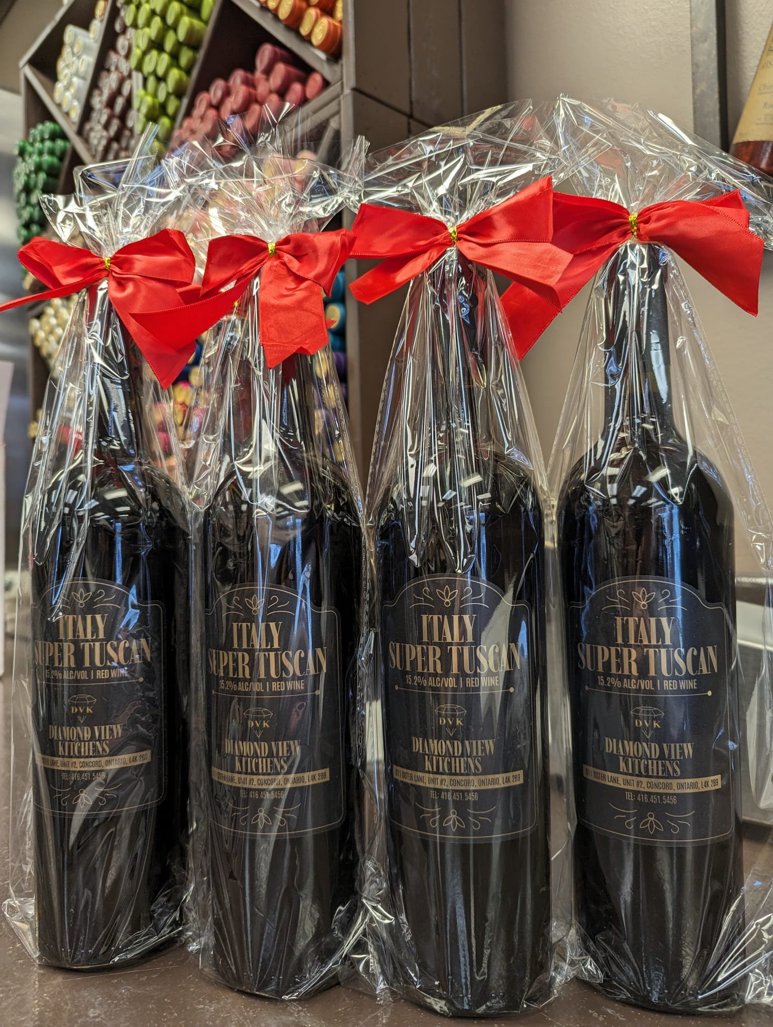 Four bottles of Italian Super Tuscan wine wrapped in clear plastic with red bows, displayed in front of a shelf filled with various colored wine corks.