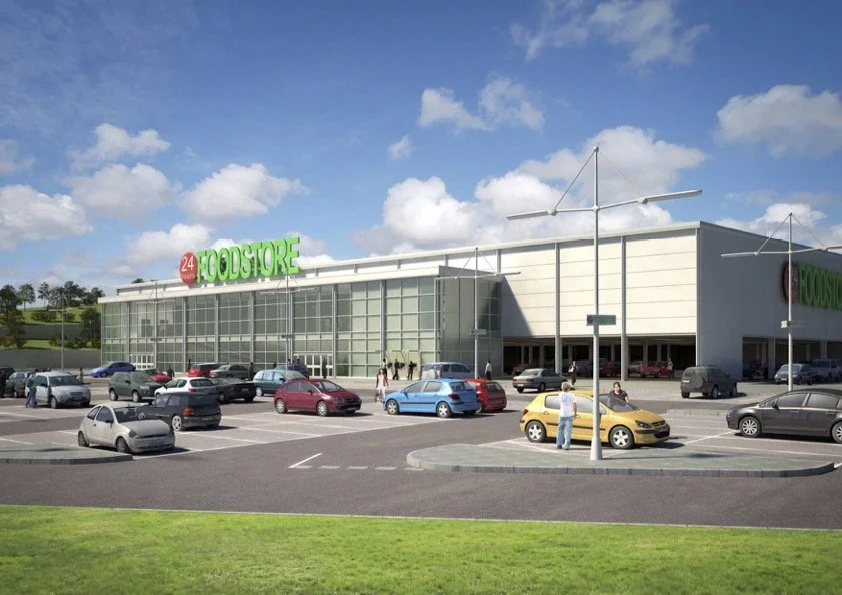 A large grocery store with a parking lot filled with cars, a few people walking, and a blue sky with clouds.