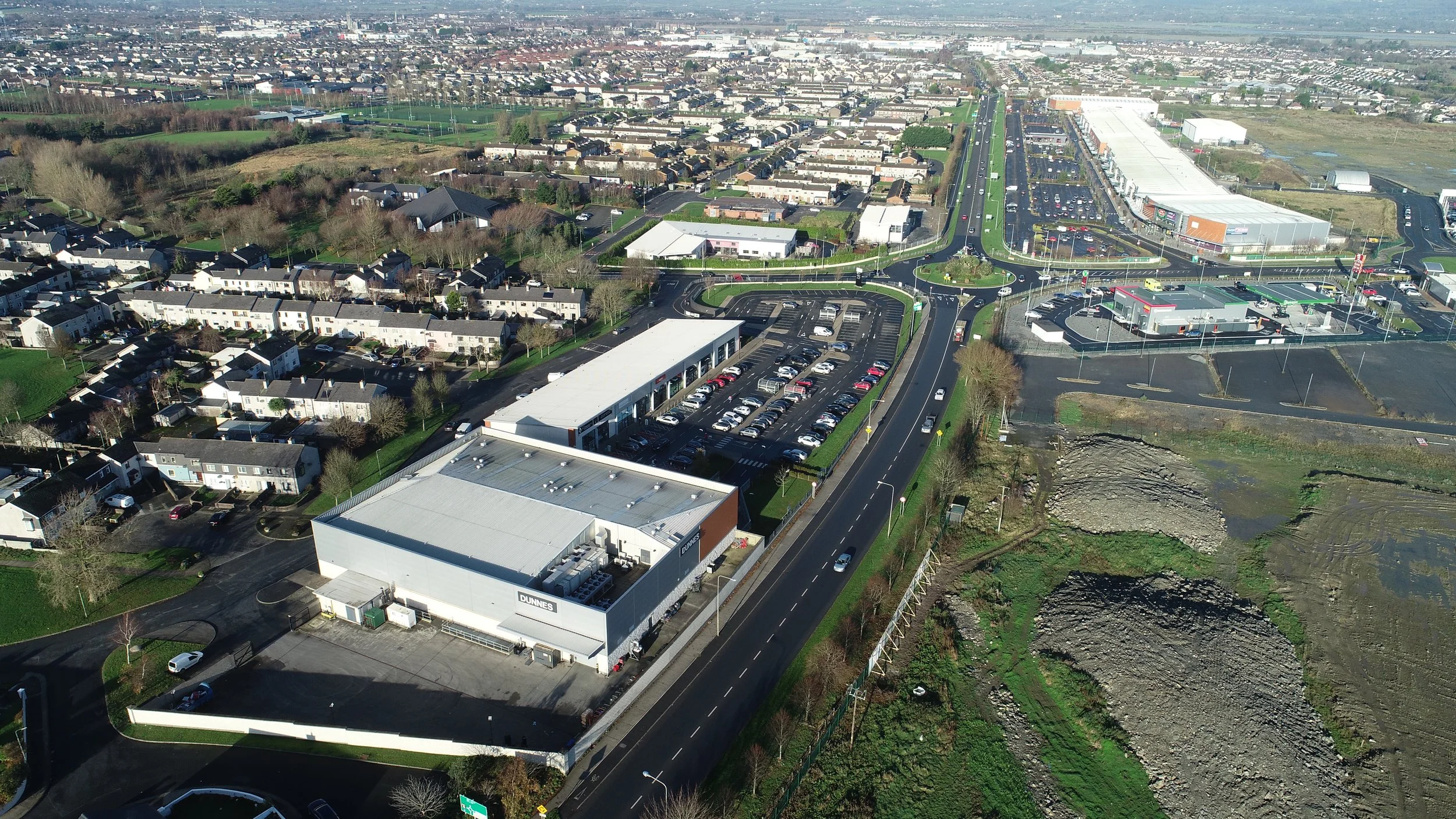 Aerial view of a suburban area showing residential houses, commercial buildings, parking lots, roads, and open land with patches of grass and dirt.