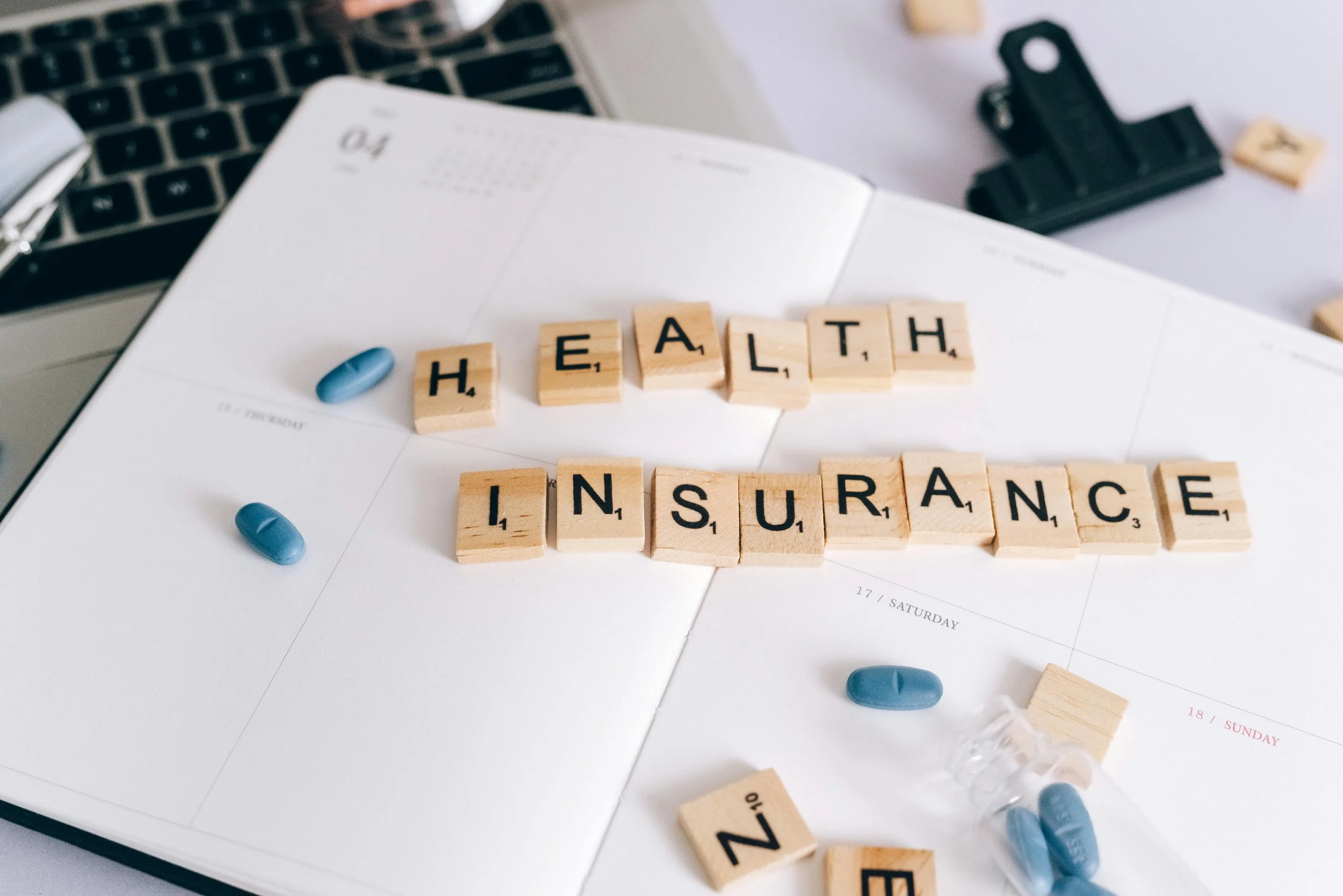 Wooden blocks spelling 'health insurance', blue pills, and a planner on a laptop keyboard.