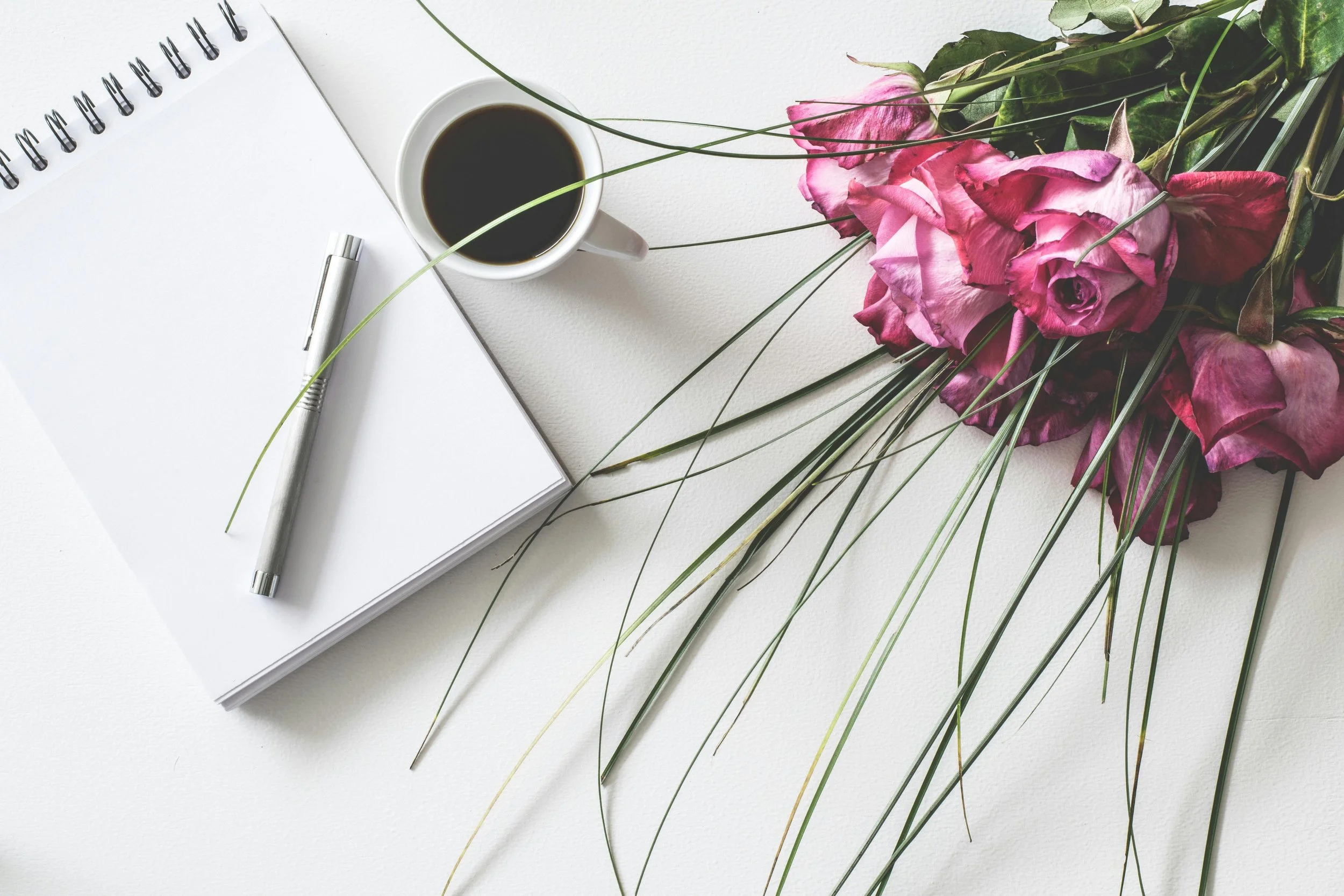 A white spiral notebook with a pen on top, a cup of black coffee, and a bouquet of pink roses with green leaves on a white surface.