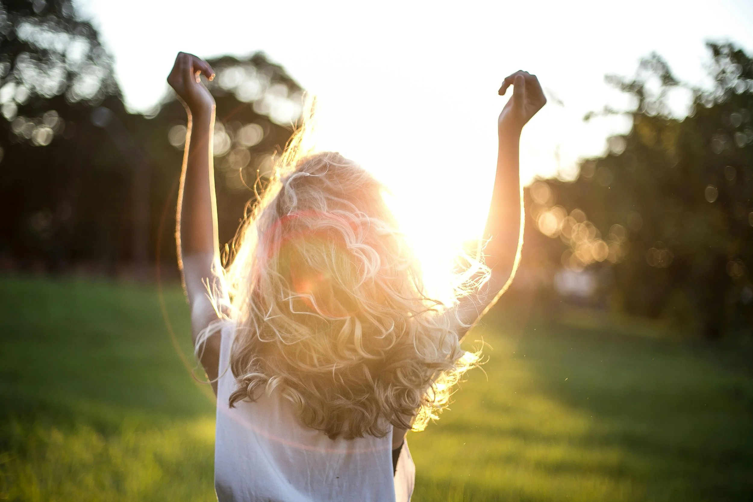 Person with blonde hair raising arms in a sunlit field.