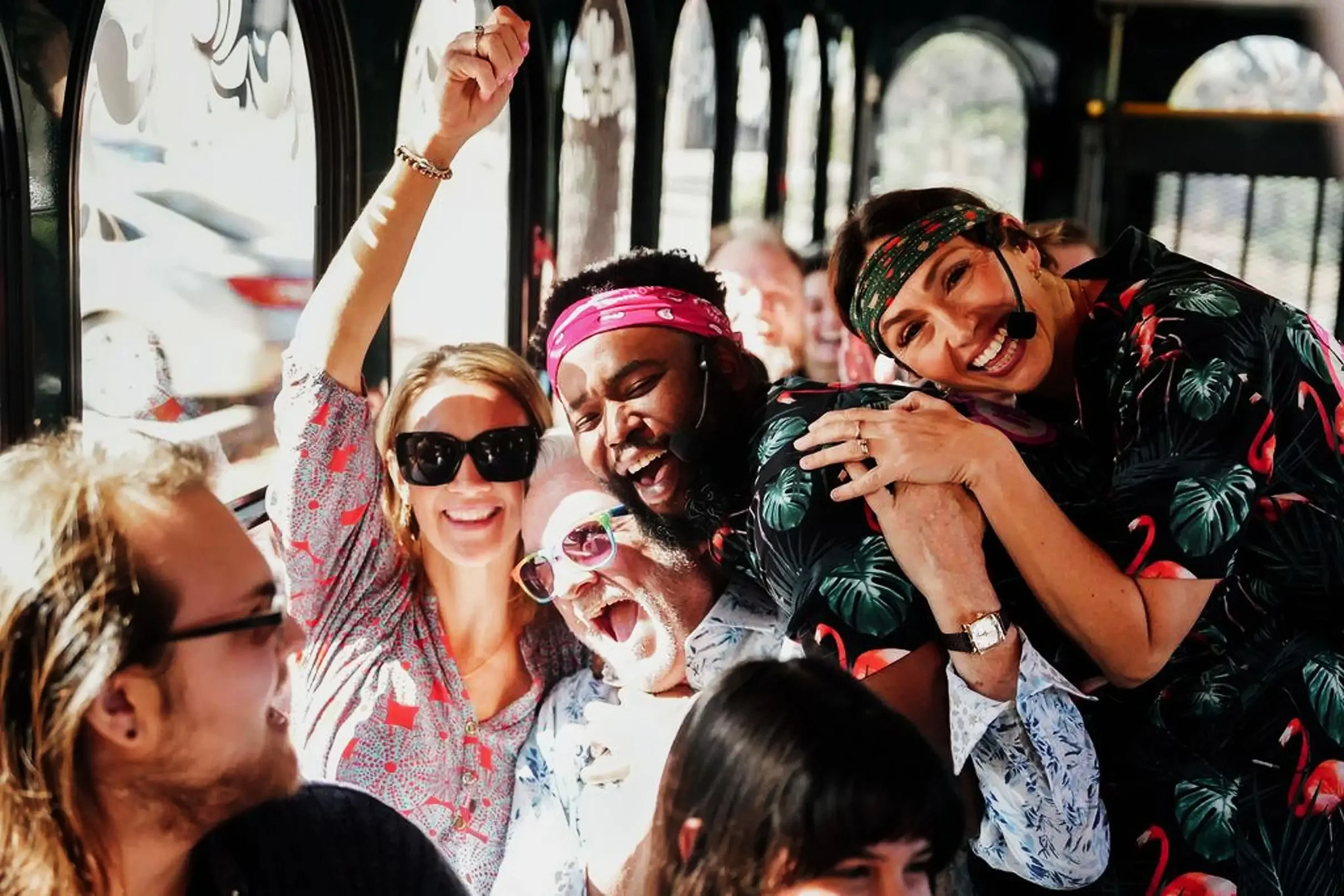 Group of diverse people celebrating and enjoying themselves on a bus, smiling, some wearing colorful headbands and sunglasses.