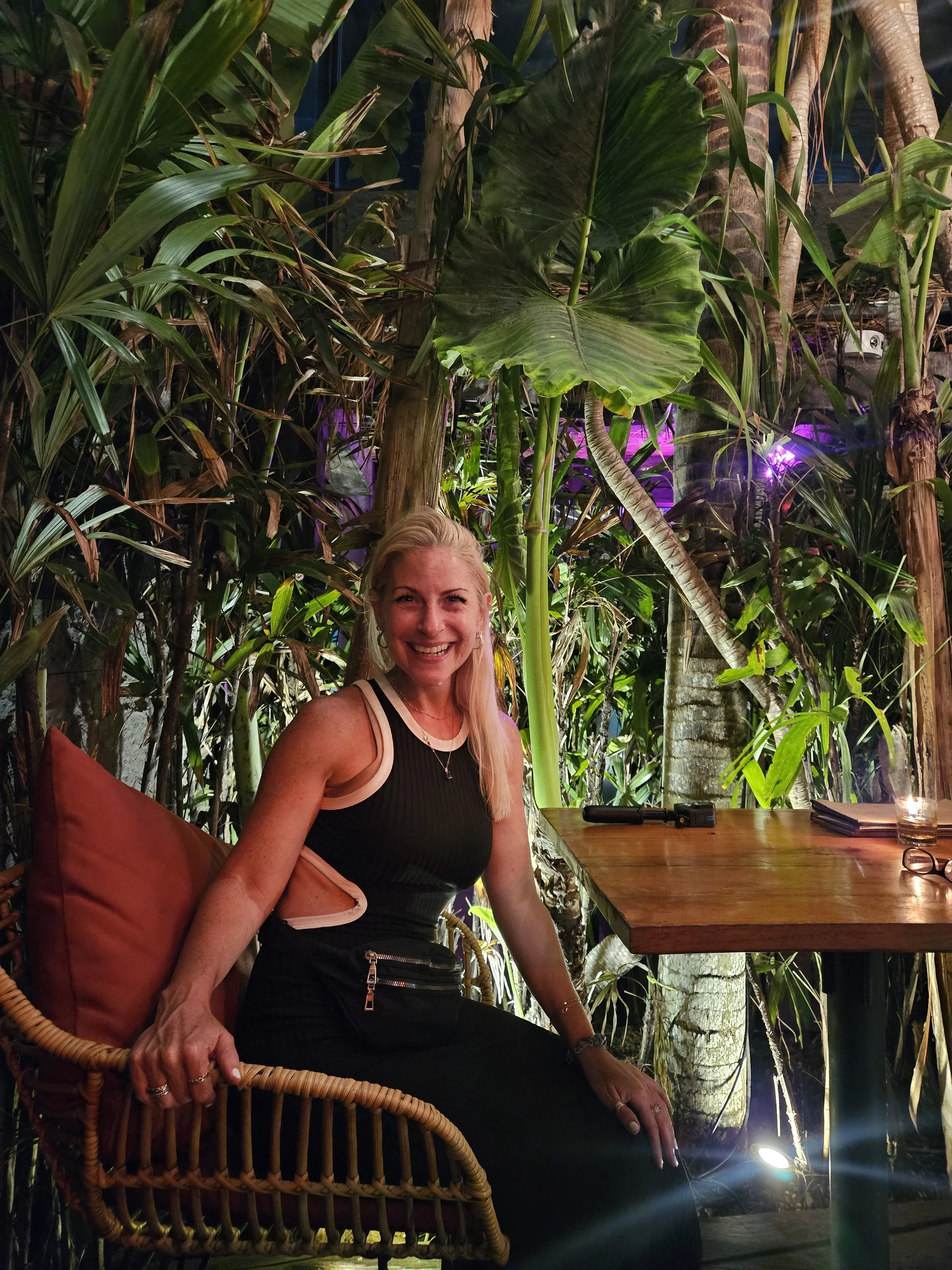 A woman with blonde hair smiling, sitting at a wooden table in a tropical setting with abundant green plants and trees.