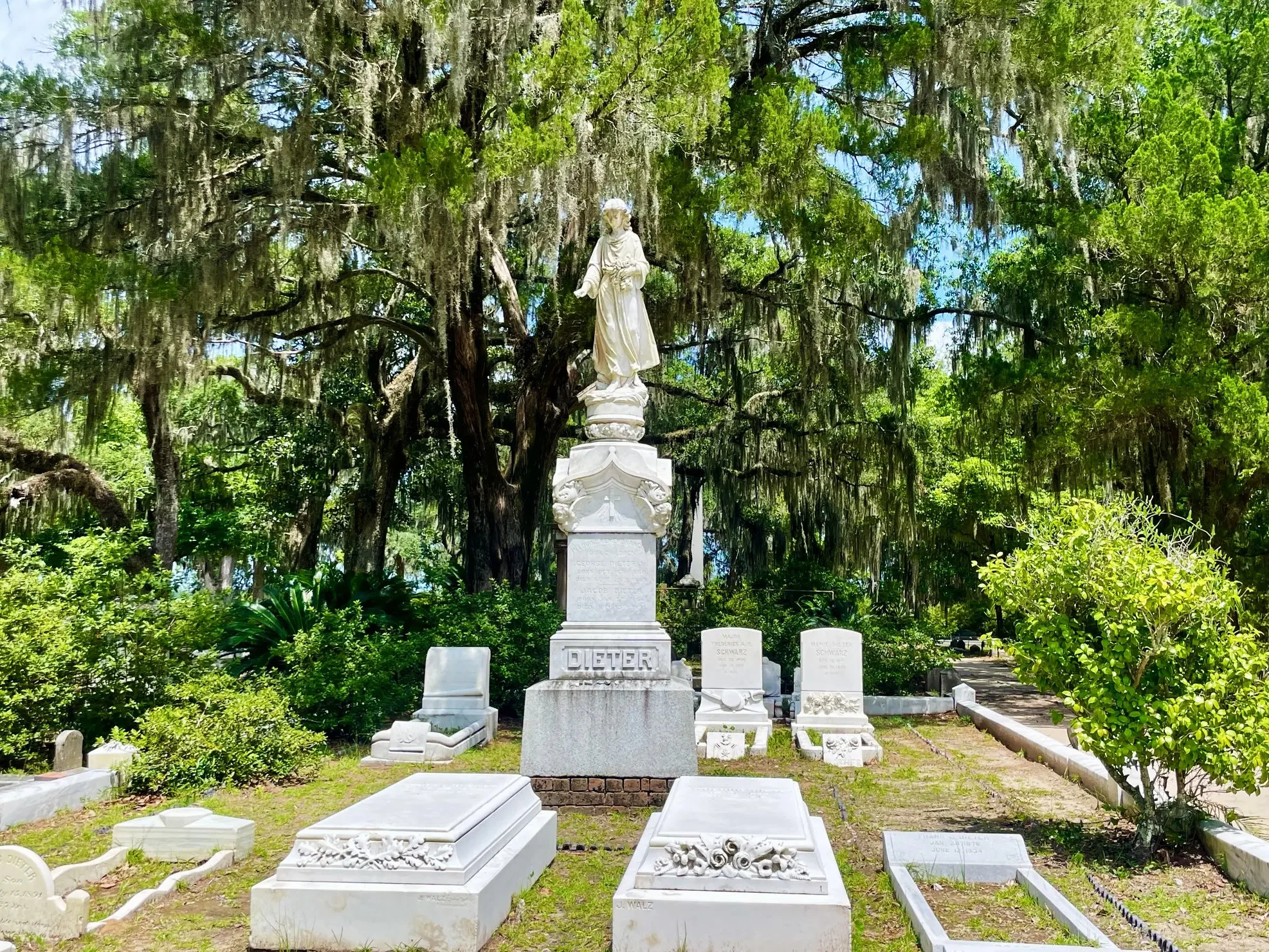 A white statue of a woman standing on a pedestal in a cemetery surrounded by small tombstones and lush green trees, with Spanish moss hanging from the branches.