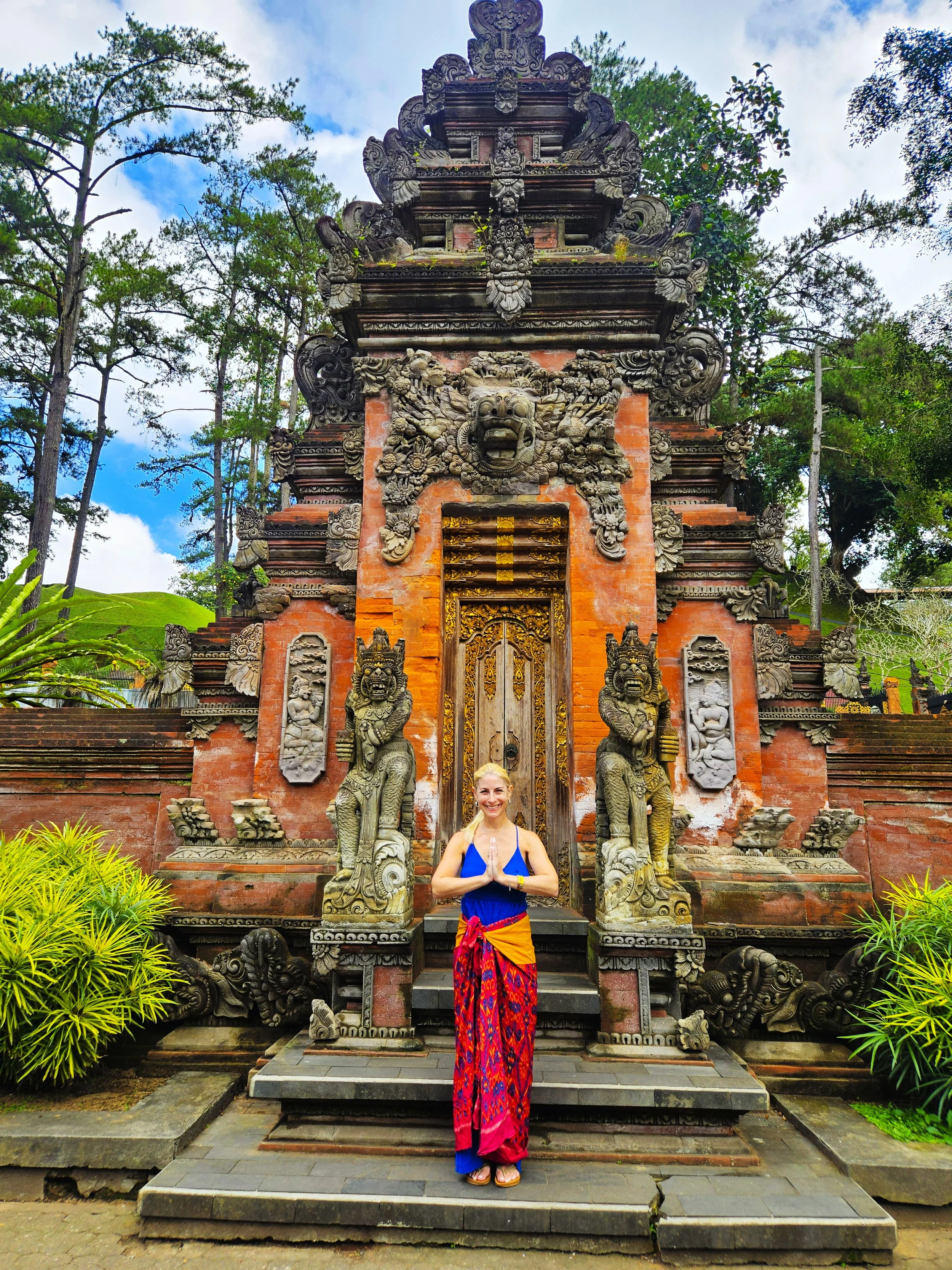A woman standing with folded hands in front of an ornate, traditional temple entrance with intricate carvings of faces and mythical creatures, surrounded by greenery and trees.