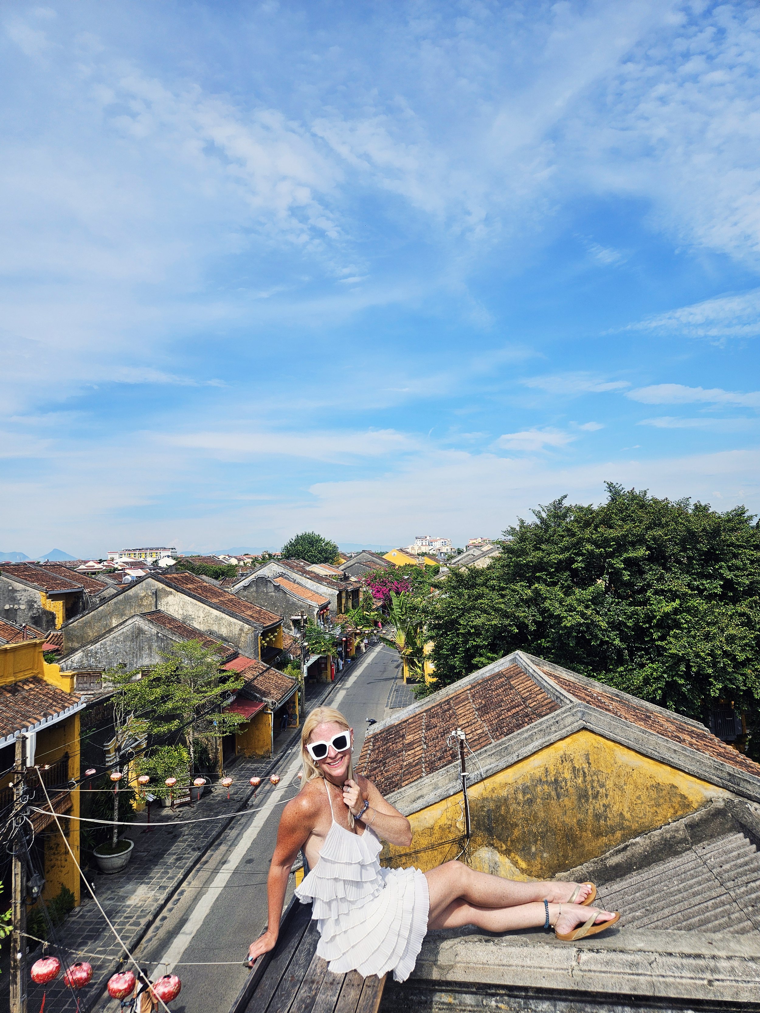 Woman with sunglasses sitting on a rooftop in a small town with orange-tiled roofs, a street below, lush greenery, and a bright blue sky with scattered clouds.
