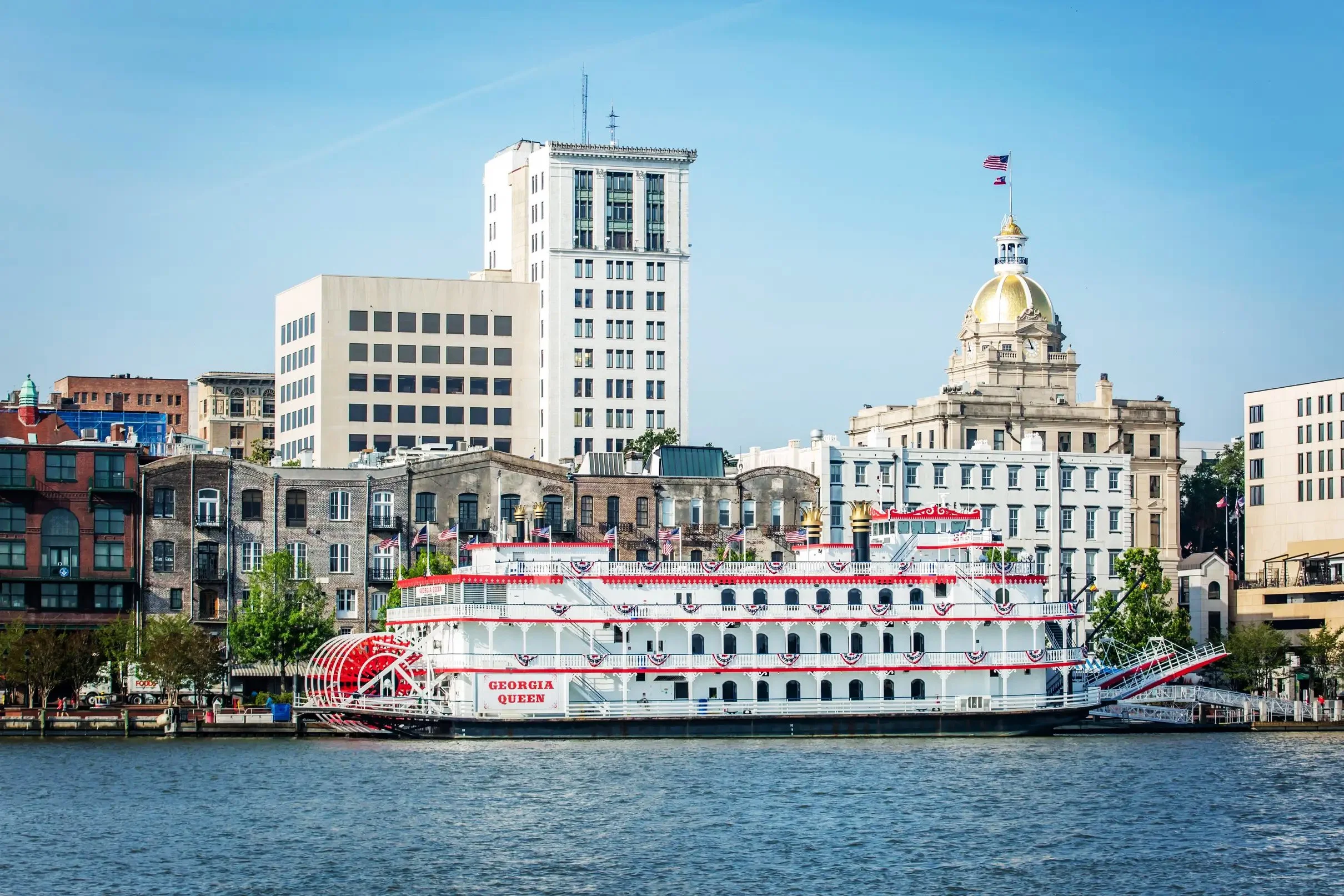 A boat docked on the river named Georgia Queen, with a city skyline in the background featuring various tall buildings including a white high-rise and a domed structure with an American flag.