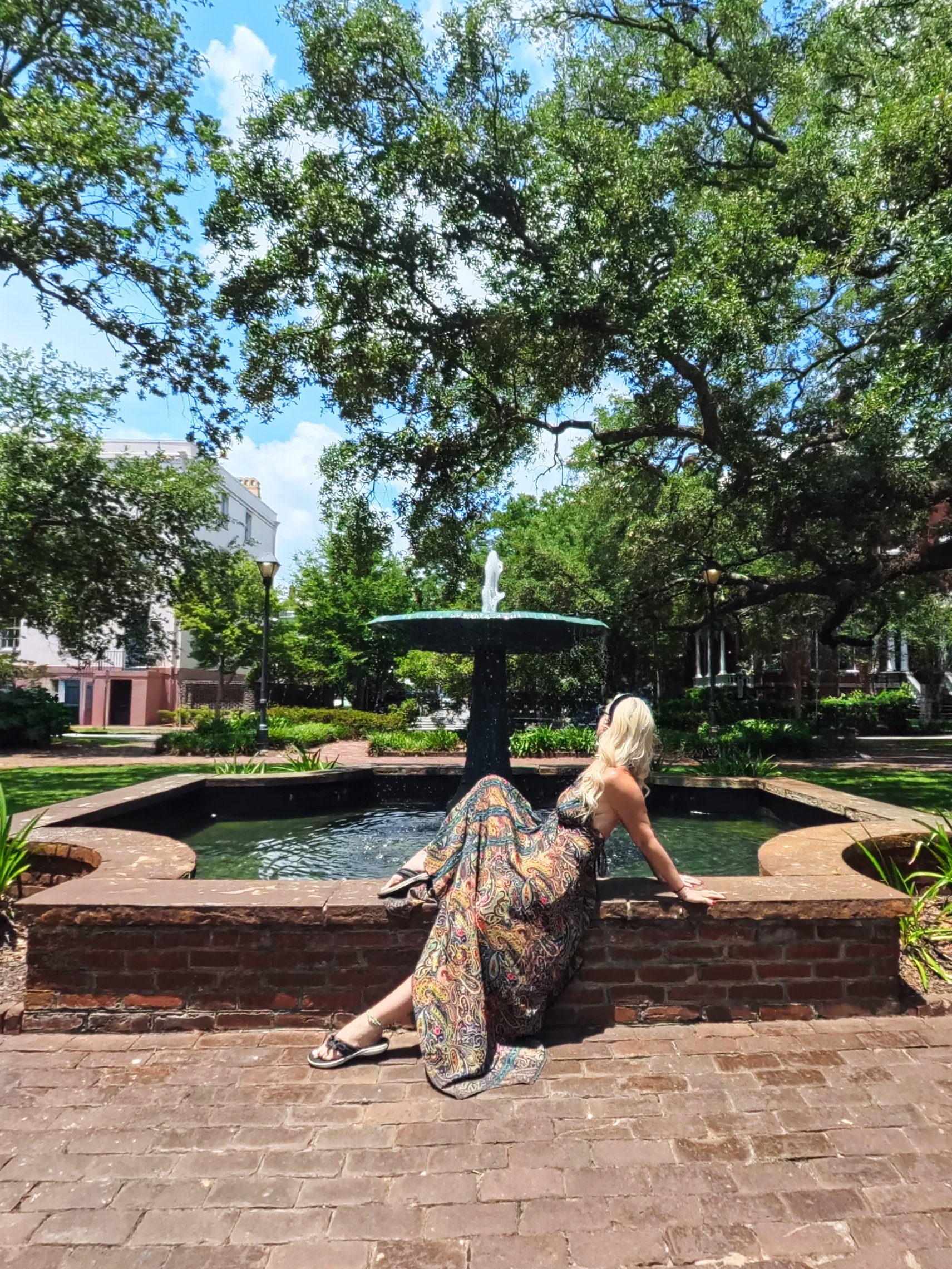 A woman with long blonde hair sitting on a brick ledge around a fountain in a park, surrounded by green trees and buildings under a blue sky with some clouds.
