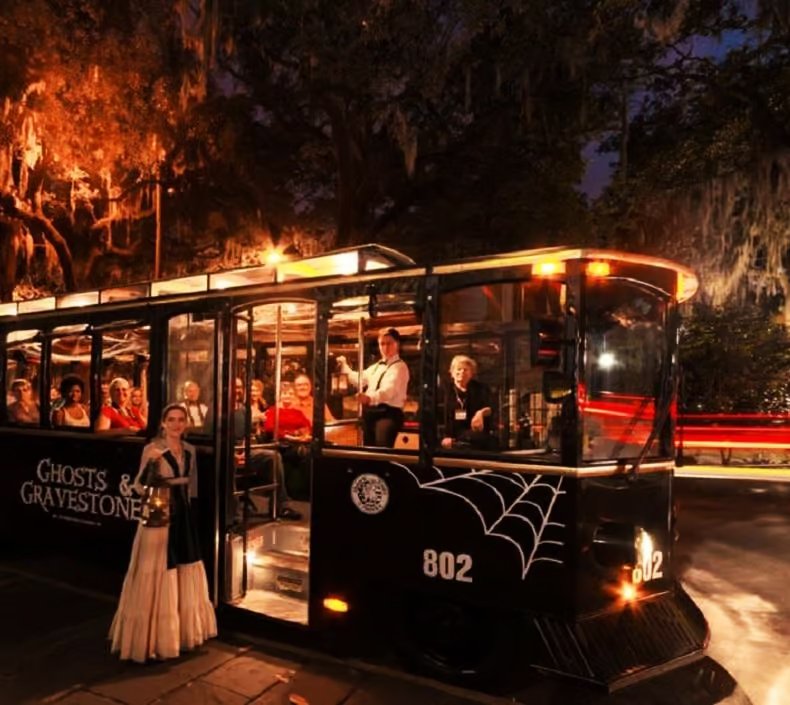 A haunted-themed trolley with ghost and spiderweb decorations, carrying several passengers at night, with people in costumes, some indoors and some waiting outside, in a spooky setting.