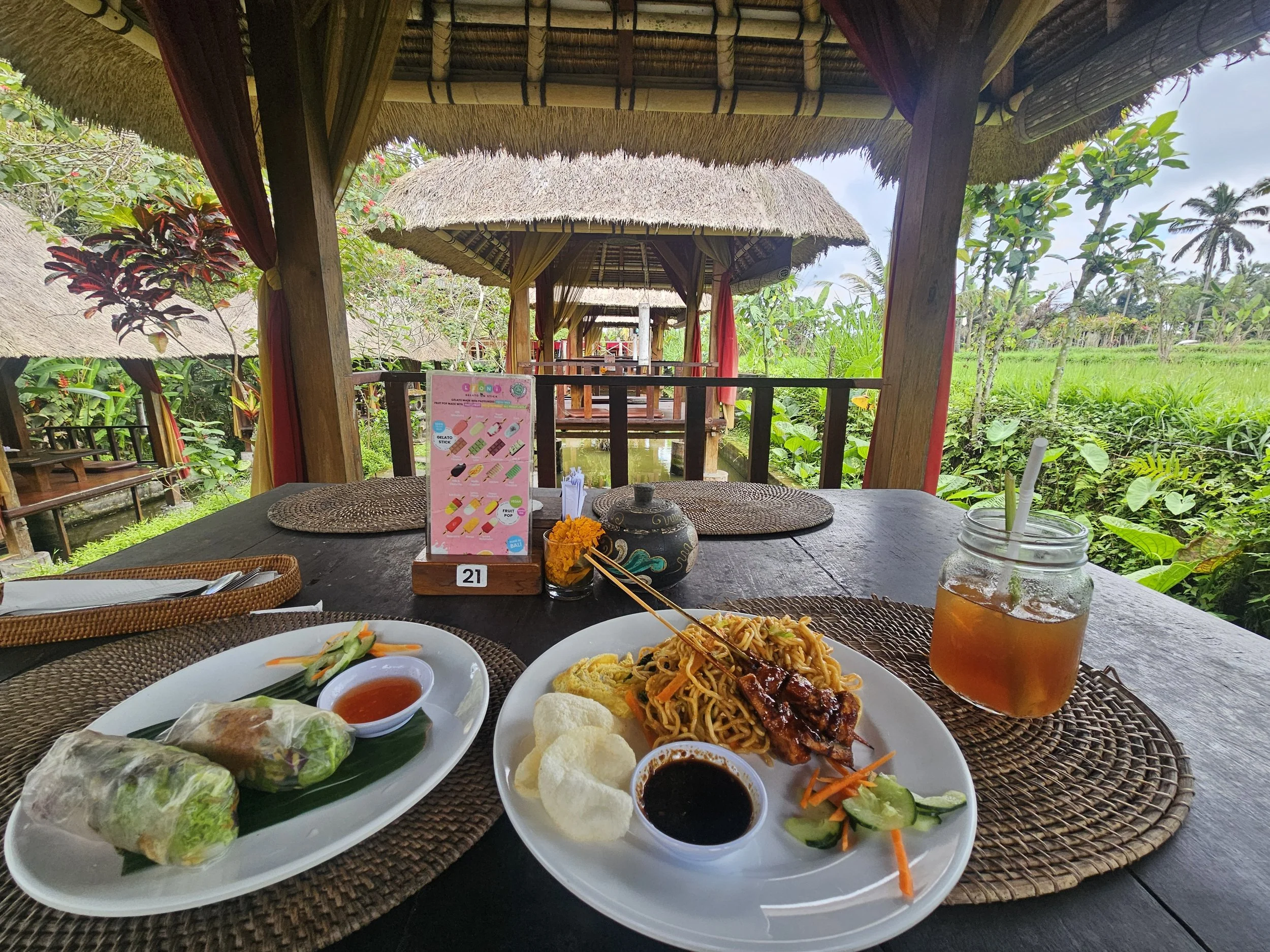 A table with several plates of Asian cuisine, including spring rolls, noodles, and dipping sauces, set in a tropical outdoor restaurant with thatched roof huts and lush green vegetation in the background.