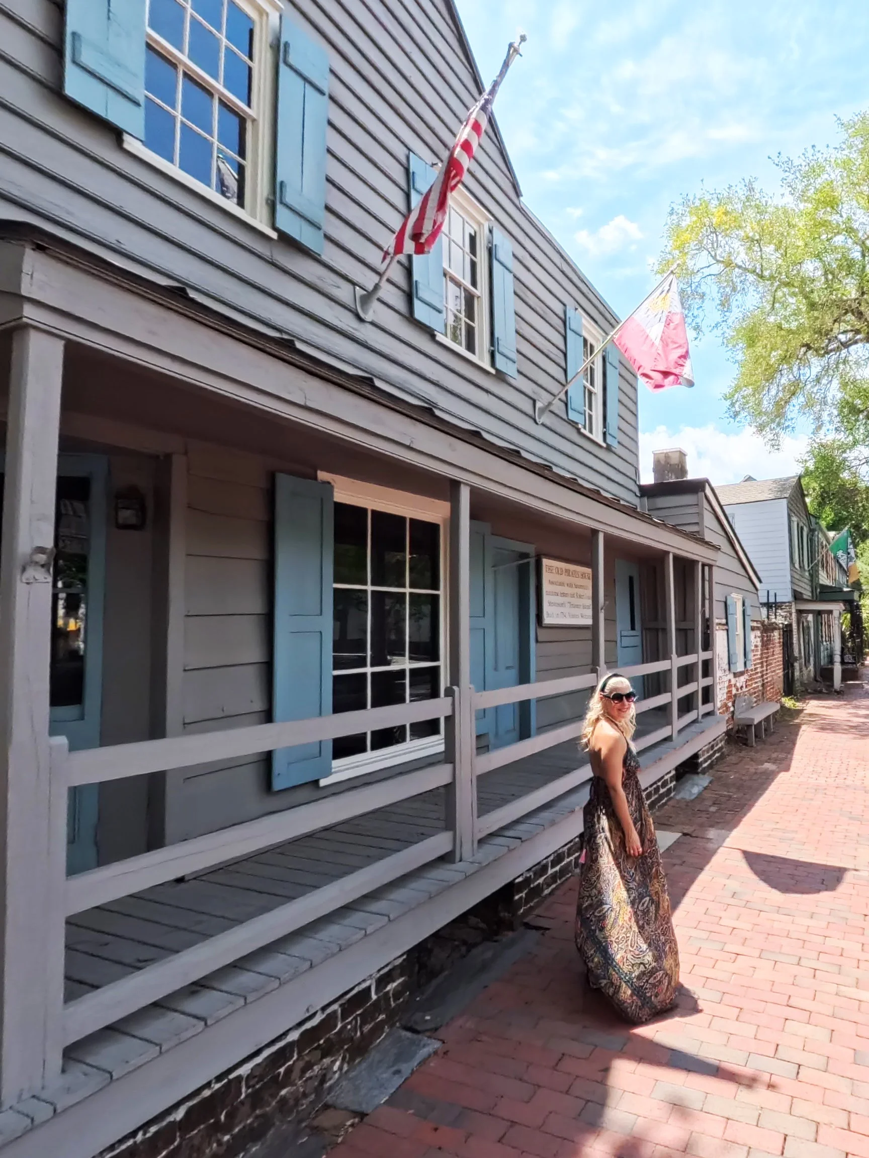 A woman in a patterned maxi dress and sunglasses standing on a brick sidewalk outside a historic building with light blue shutters and American flags flying above. The sky is blue with some clouds, and there are trees in the background.