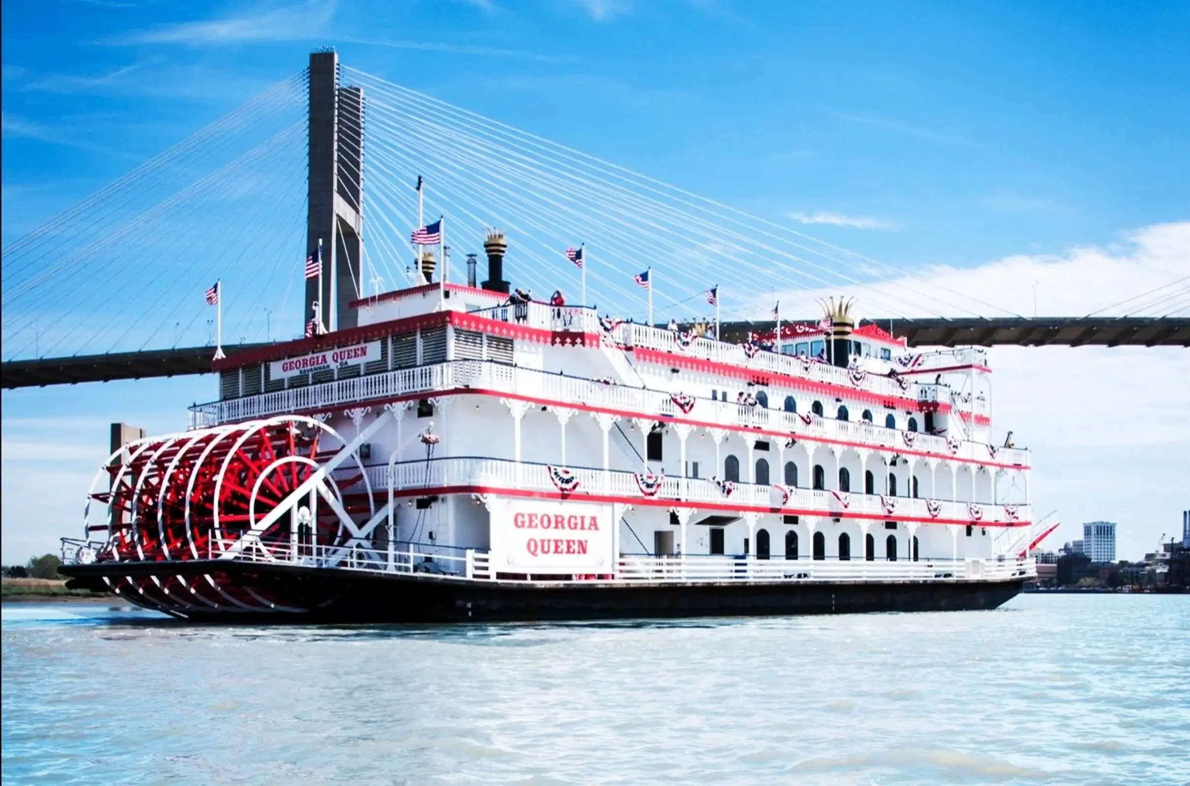 A large white paddle steamship named Georgia Queen decorated for Independence Day with red, white, and blue banners and American flags, floating on the water against a blue sky.