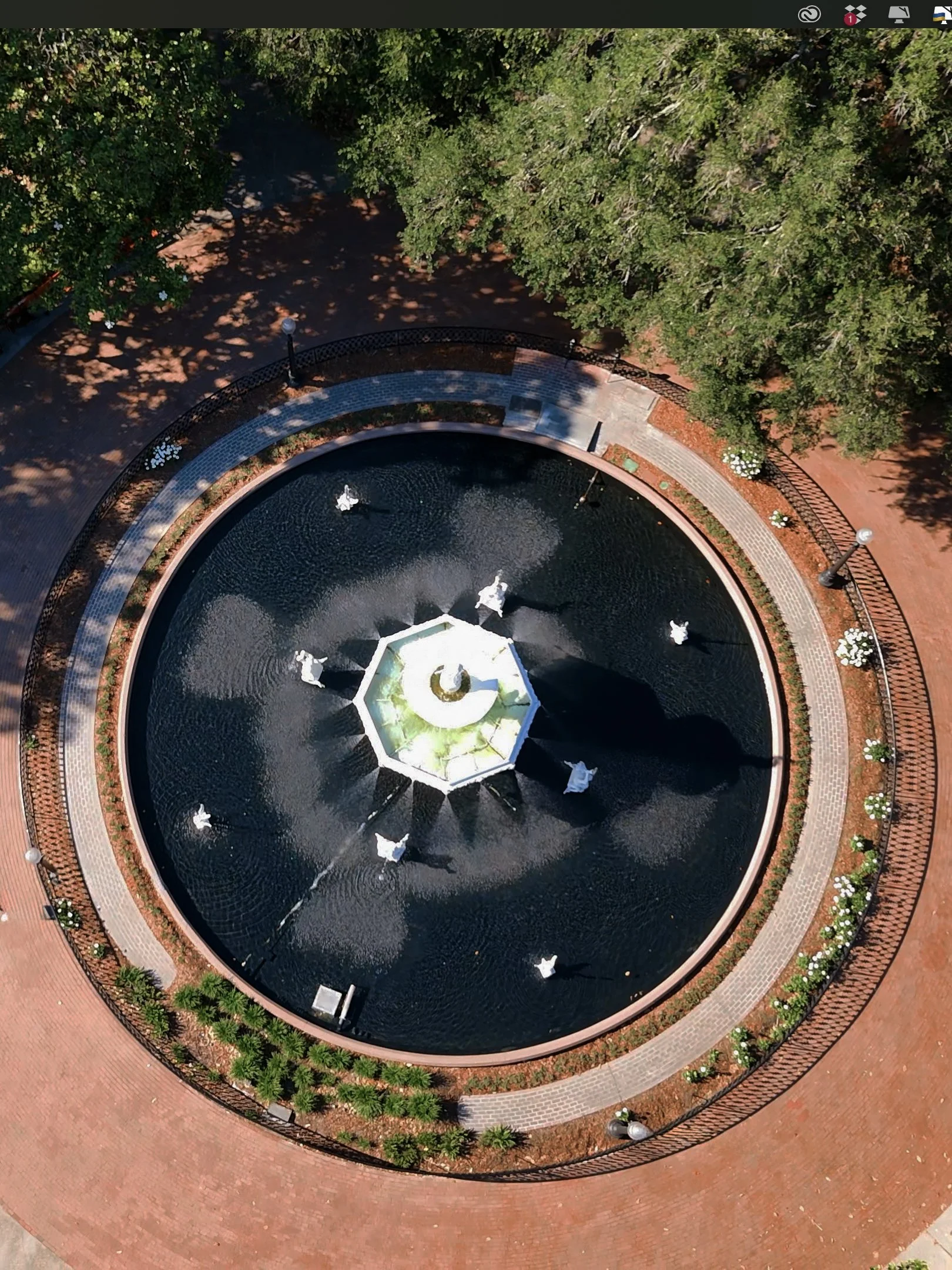 Bird's eye view of a circular fountain with sculptures surrounded by brick pathway, trees, and garden lights.