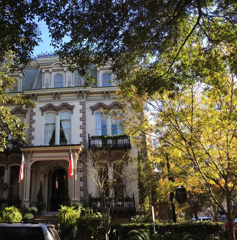 A large, historic building with Victorian architecture, with a cream-colored facade, decorative moldings, and balcony railings, partially obscured by trees with autumn foliage.