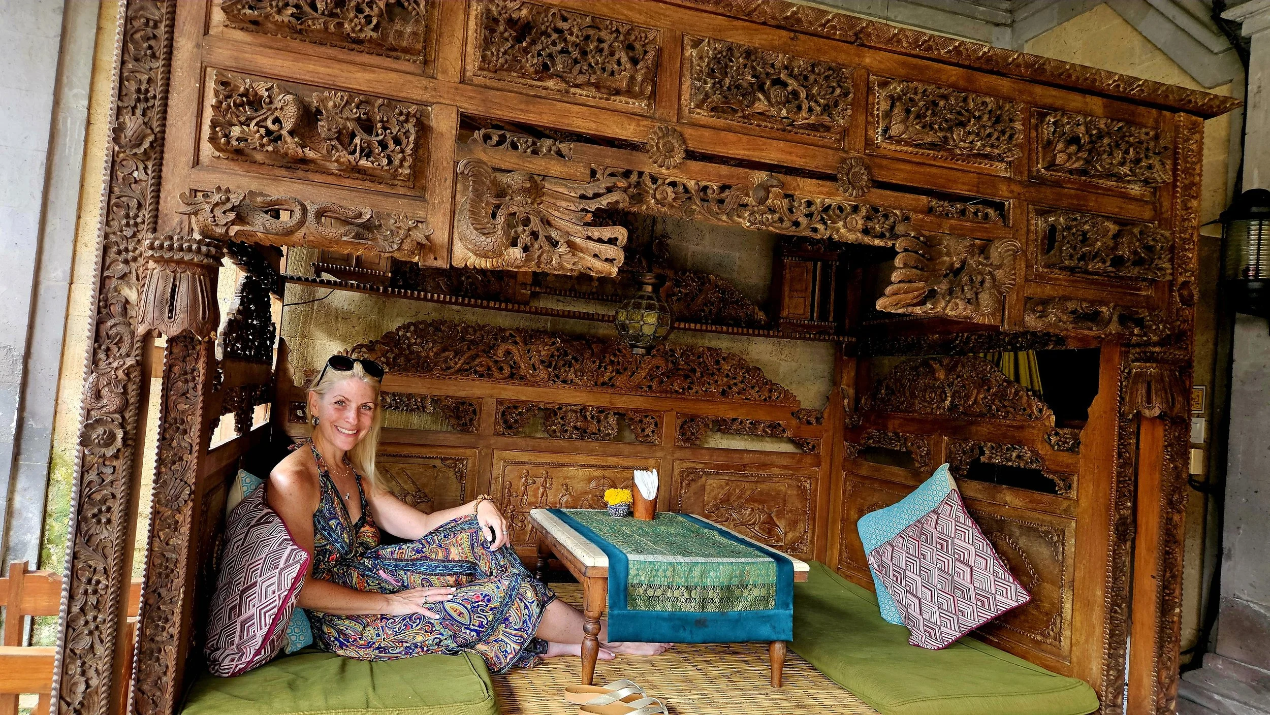 A woman sitting inside an elaborately carved wooden structure with intricate floral and dragon designs, smiling at the camera, with colorful cushions and a small table nearby.
