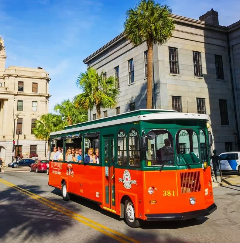 A vintage red and green trolley bus filled with passengers, traveling on a city street lined with palm trees and historic buildings.