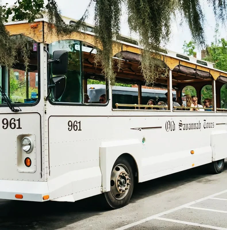 Vintage white trolley bus with open-air seating, labeled 'Old Savannah Tours,' parked on the street amidst trees.