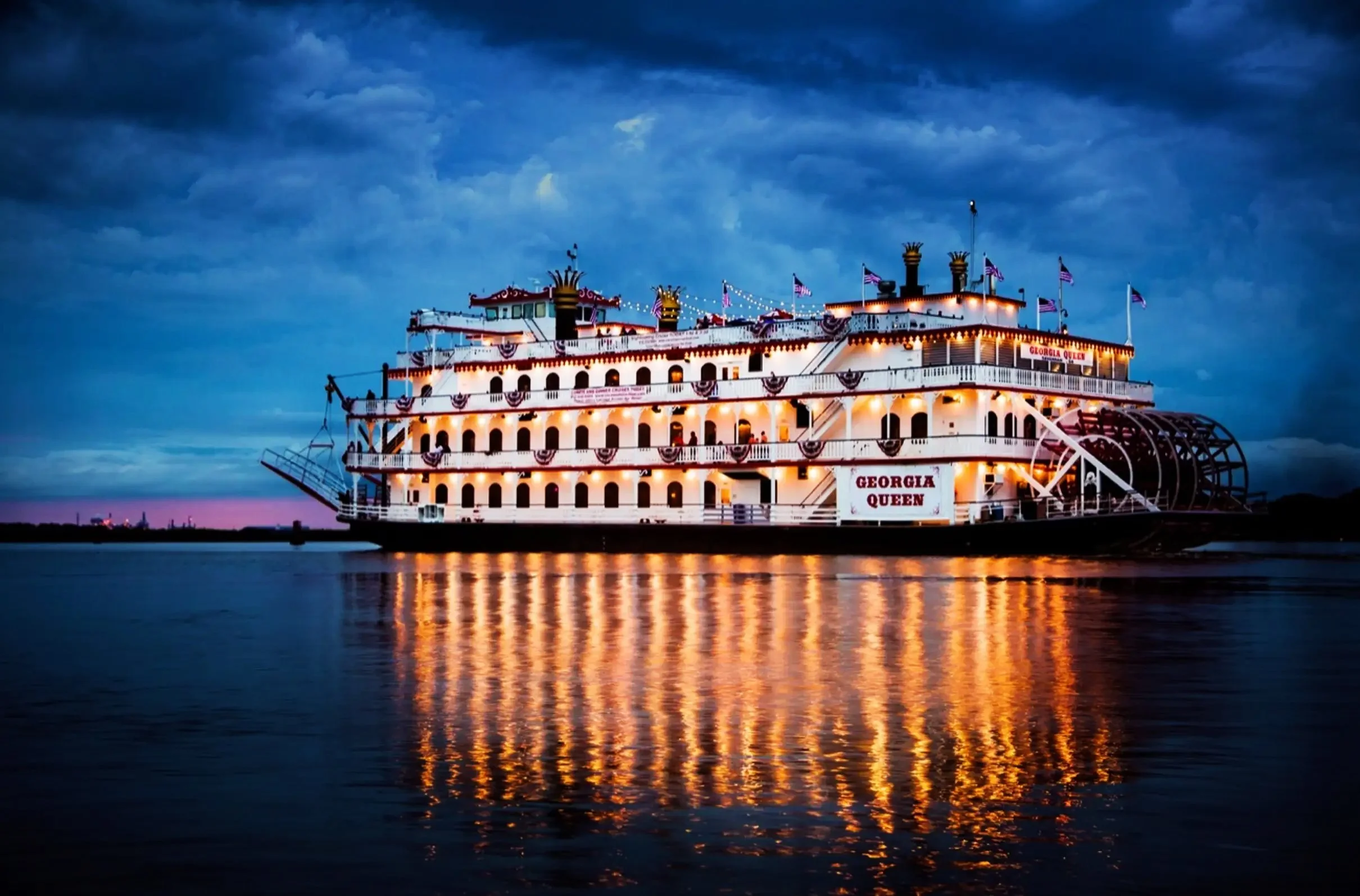 A large, illuminated paddle wheel riverboat named Georgia Queen sailing on a calm water surface during dusk, with dark clouds in the sky and reflections of the boat's lights on the water.