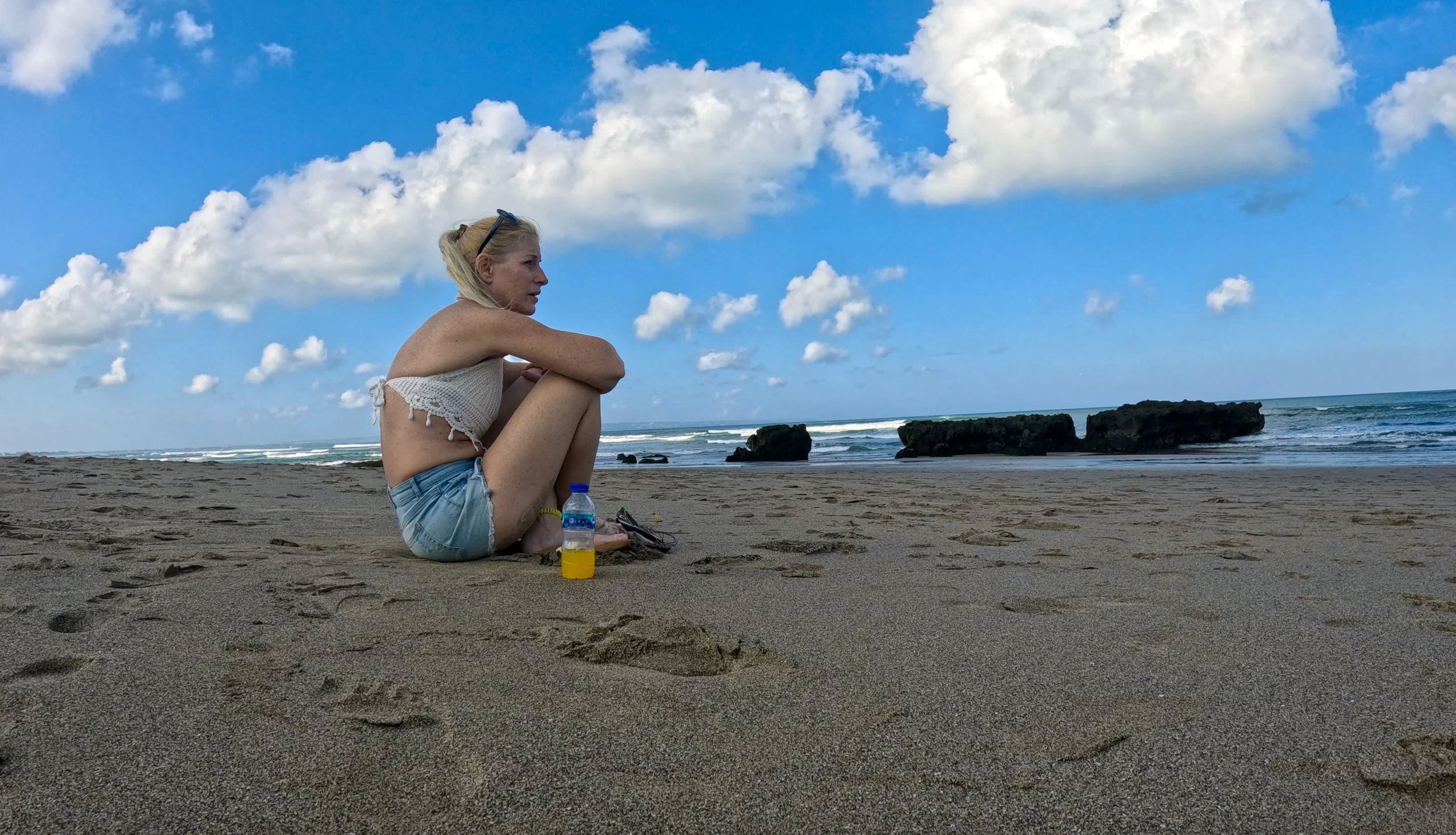 A woman sitting on the sandy beach with her knees up, looking out at the ocean and blue sky. She wears a white crochet top, denim shorts, and sunglasses. There is a water bottle and some personal items on the sand nearby, with rocky formations and gentle waves in the background.