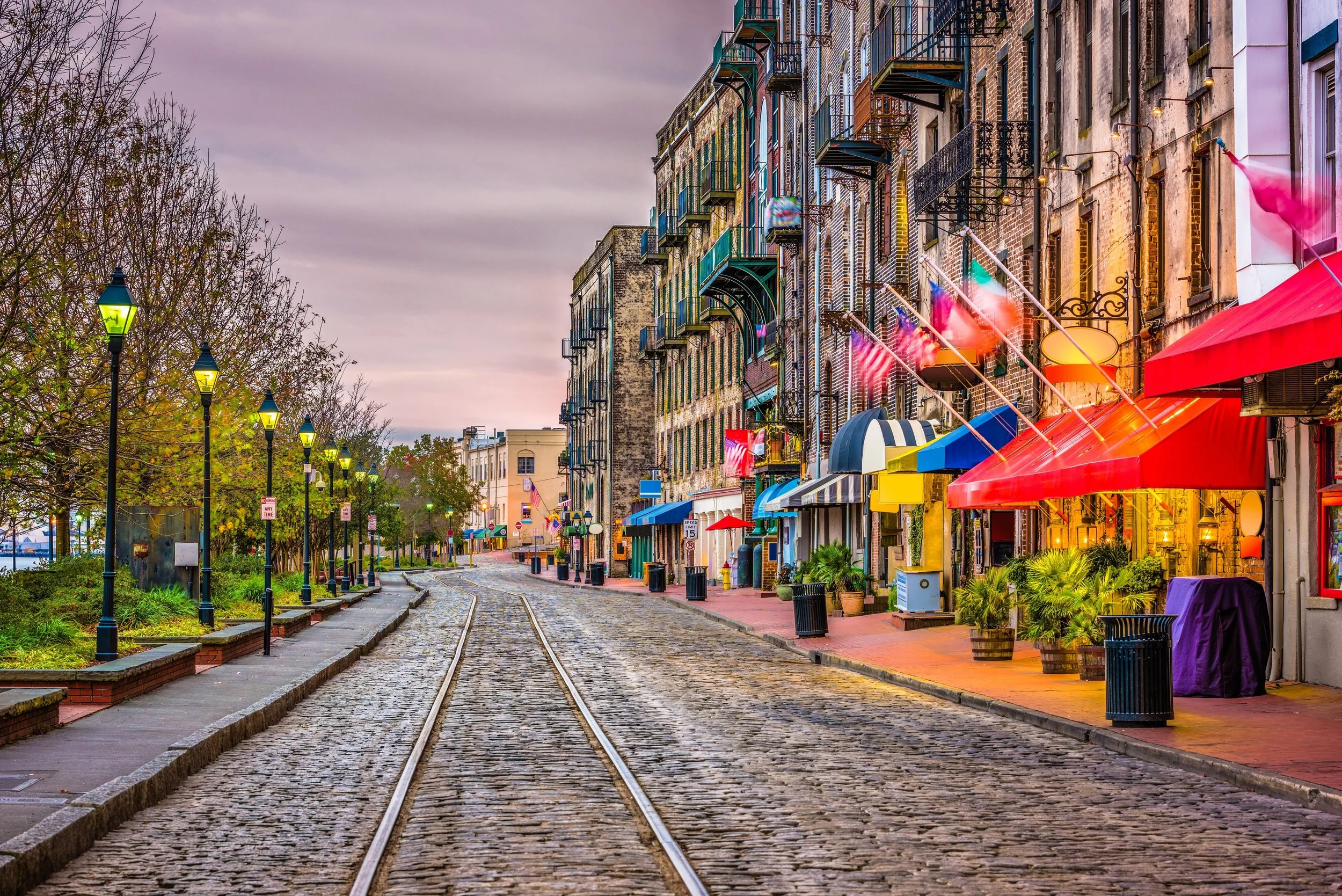 A cobblestone street with tram tracks, lined with colorful buildings with fire escape balconies and brightly lit shopfronts, during dusk with a cloudy sky.
