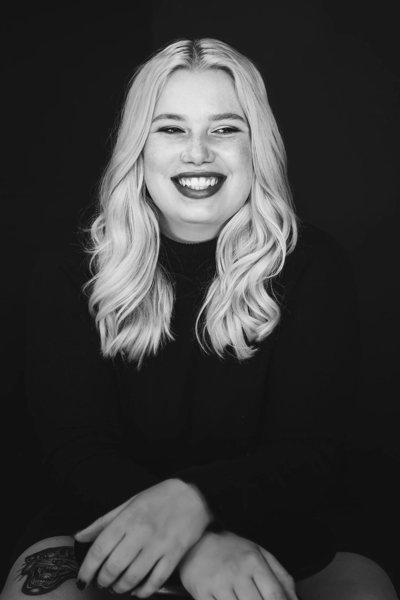 Portret van een lachende vrouw met lang, golvend blond haar, een neuspiercing en een tattoo op haar linkerhand, gekleed in een zwarte trui, tegen een zwarte achtergrond.