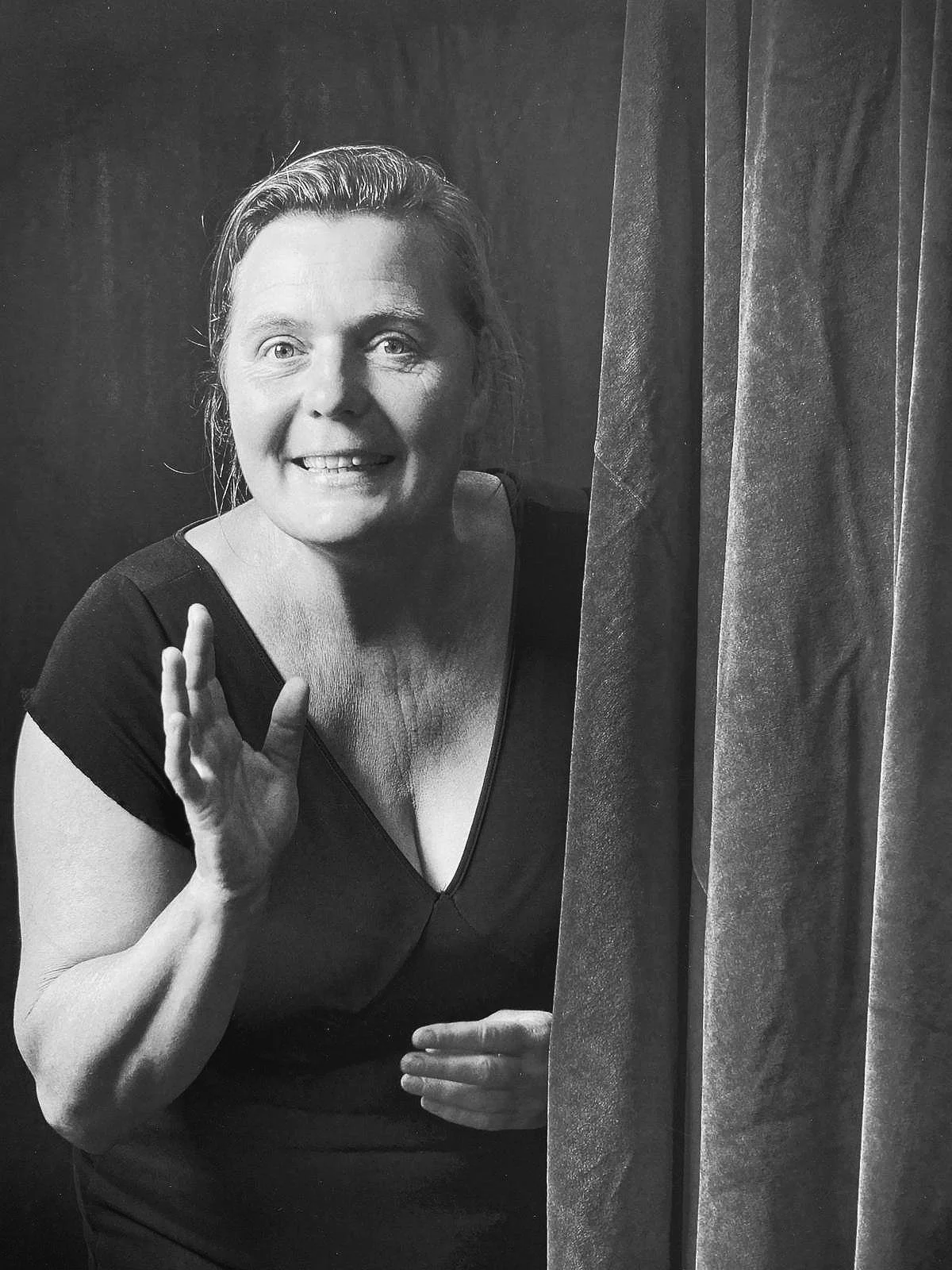 Een zwart-wit foto van een glimlachende vrouw die uit een gordijn vandaan komt, haar hand omhoog houdt en oogcontact maakt.