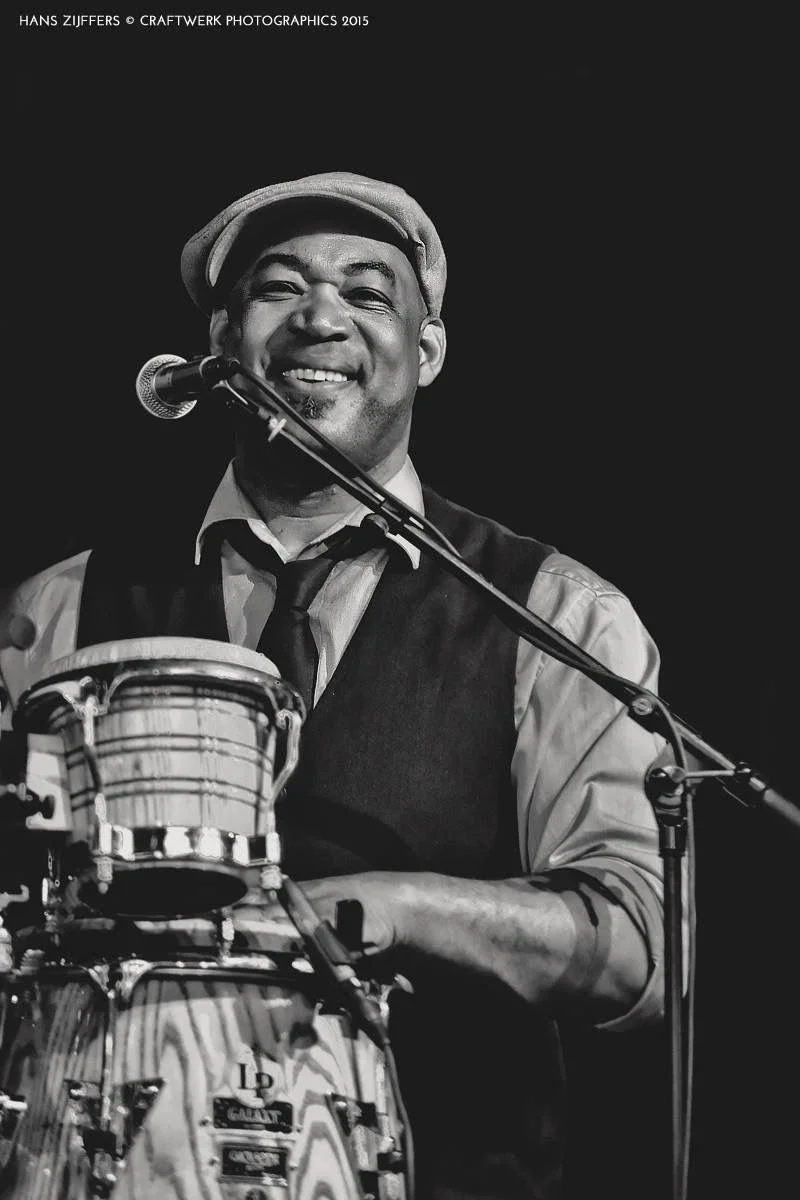 Zwart-witfoto van een glimlachende man die op een drum speelt en een microfoon vasthoudt, met een hoed en een vest, op een podium.