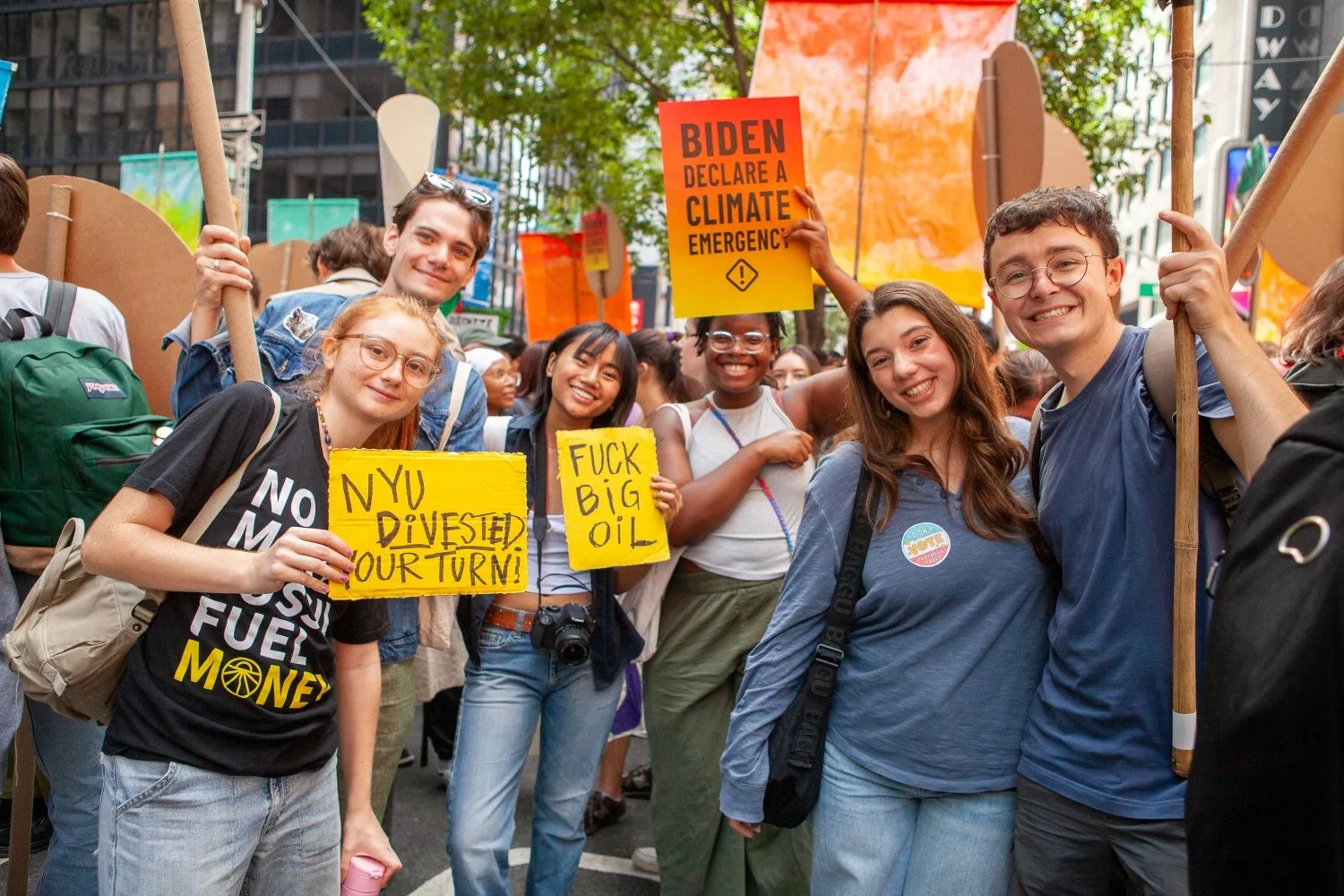Group of diverse young protesters smiling and holding signs at a climate rally in an urban setting.