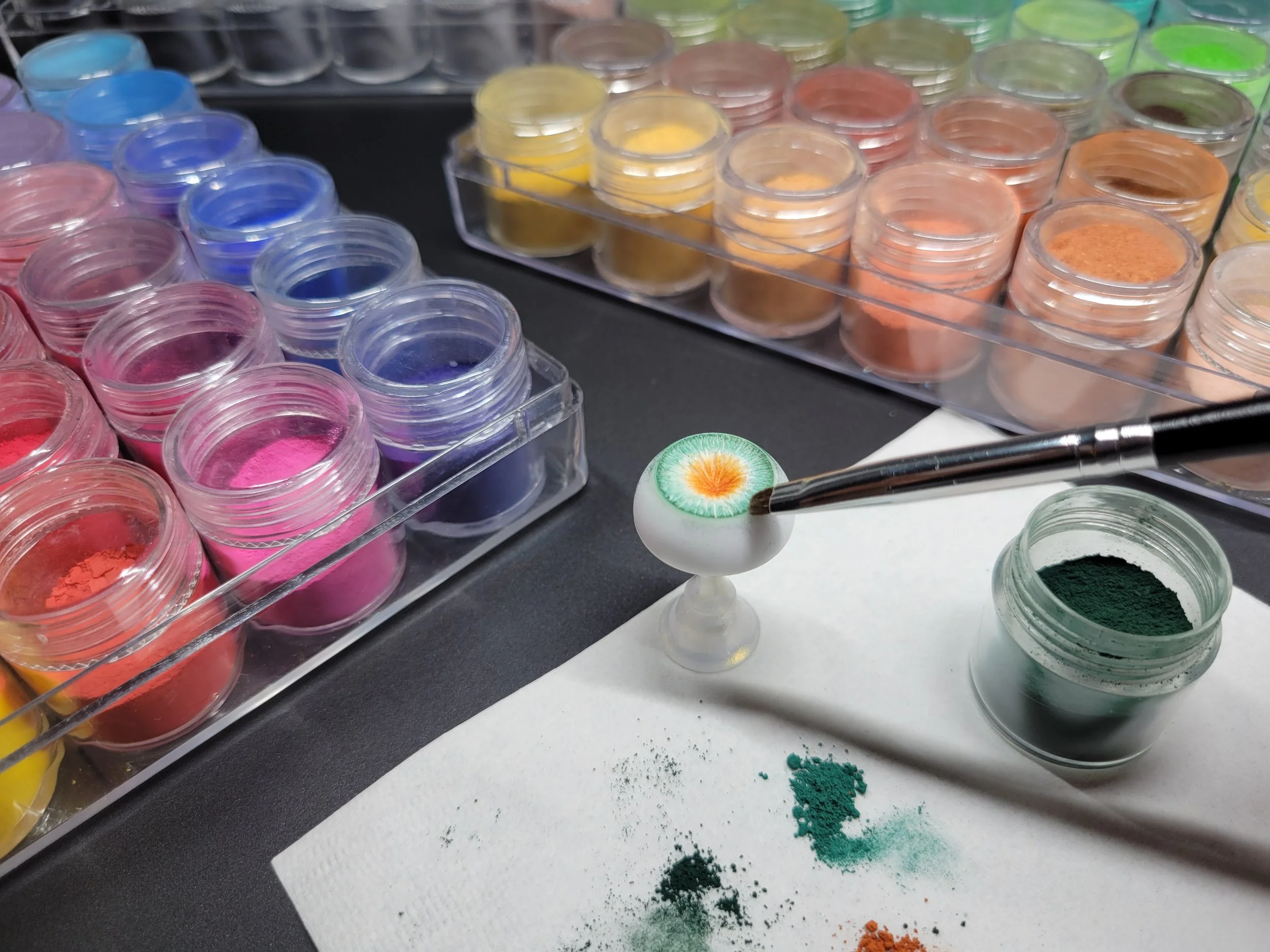 Close-up of small containers filled with colorful pigments arranged in rows on a black surface with spilled pigment powders and a brush nearby.