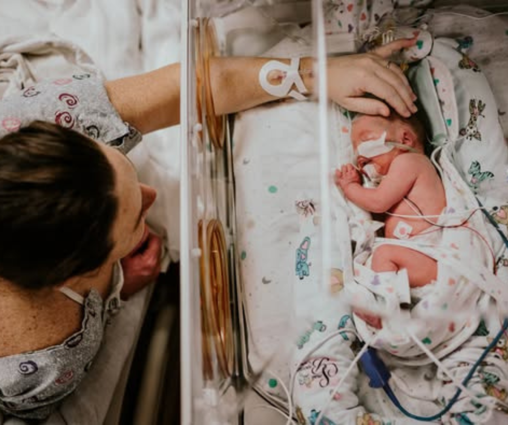 Mother leaning over a neonatal incubator, looking at her newborn baby who is connected to medical tubes and monitors in a hospital pediatric unit.